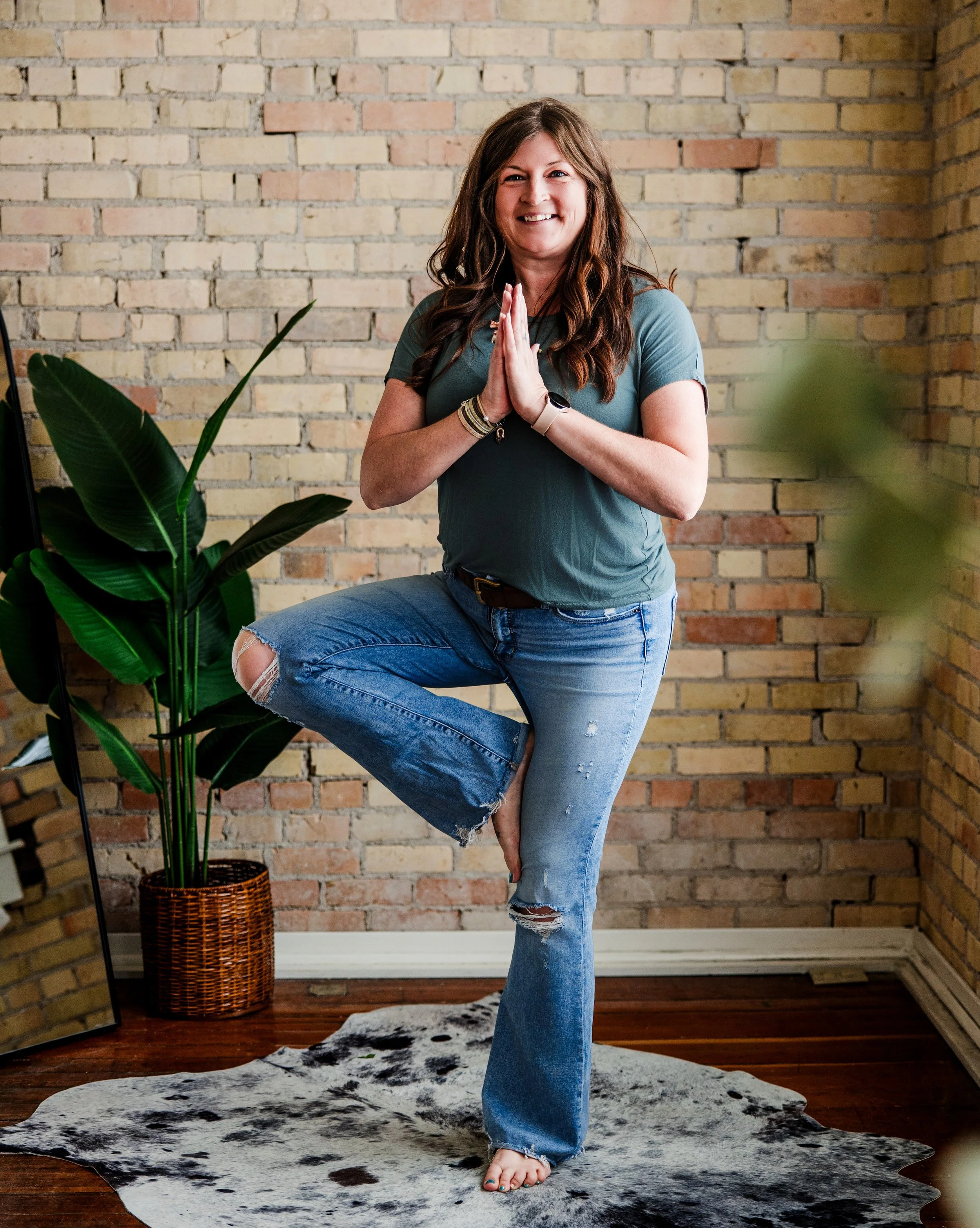 Woman in relaxed pose with one leg up, standing barefoot on a cowhide rug in front of a brick wall, with a plant in a basket to her left.