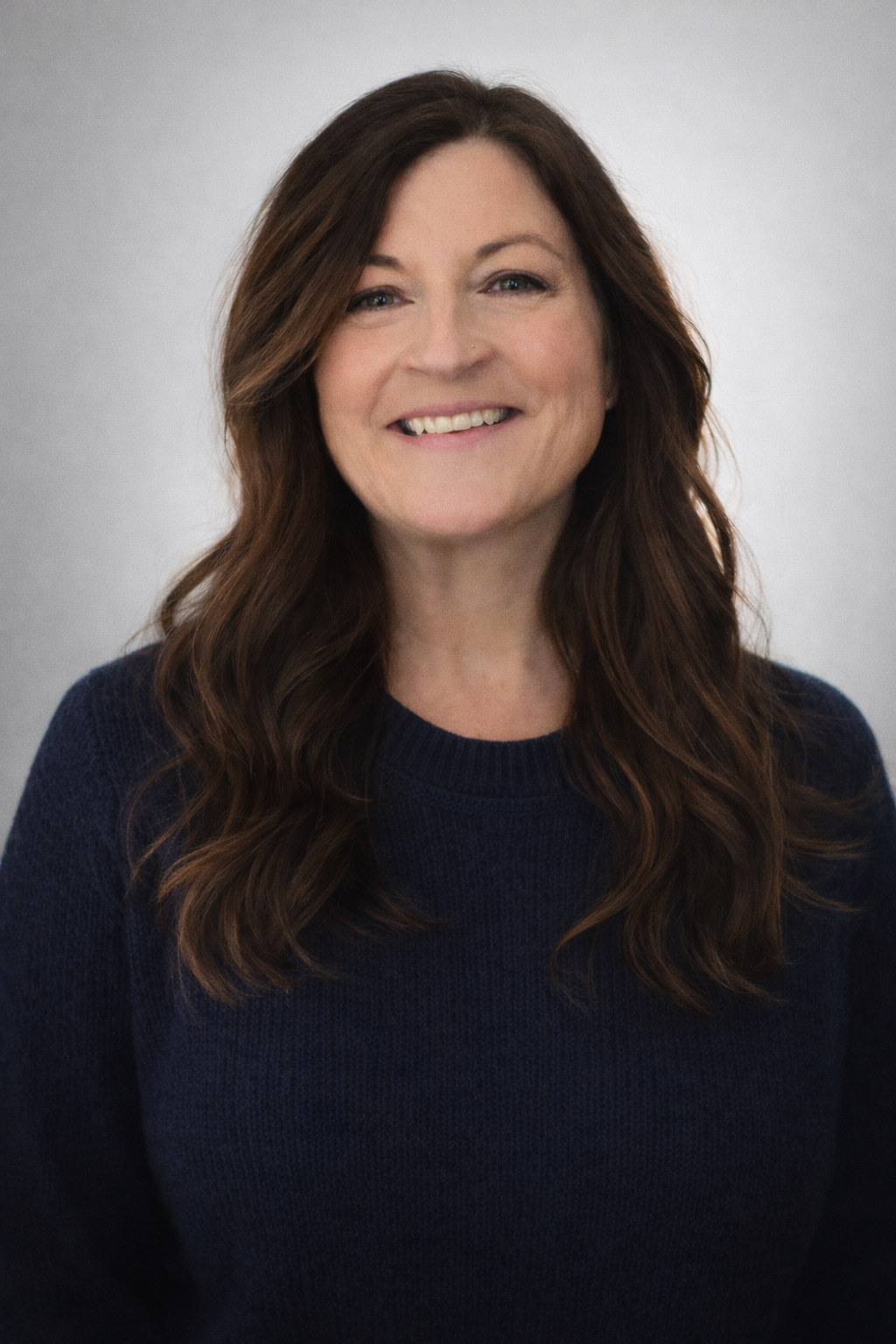 A smiling mental health therapist with long, wavy brown hair wearing a dark blue sweater against a plain gray background.