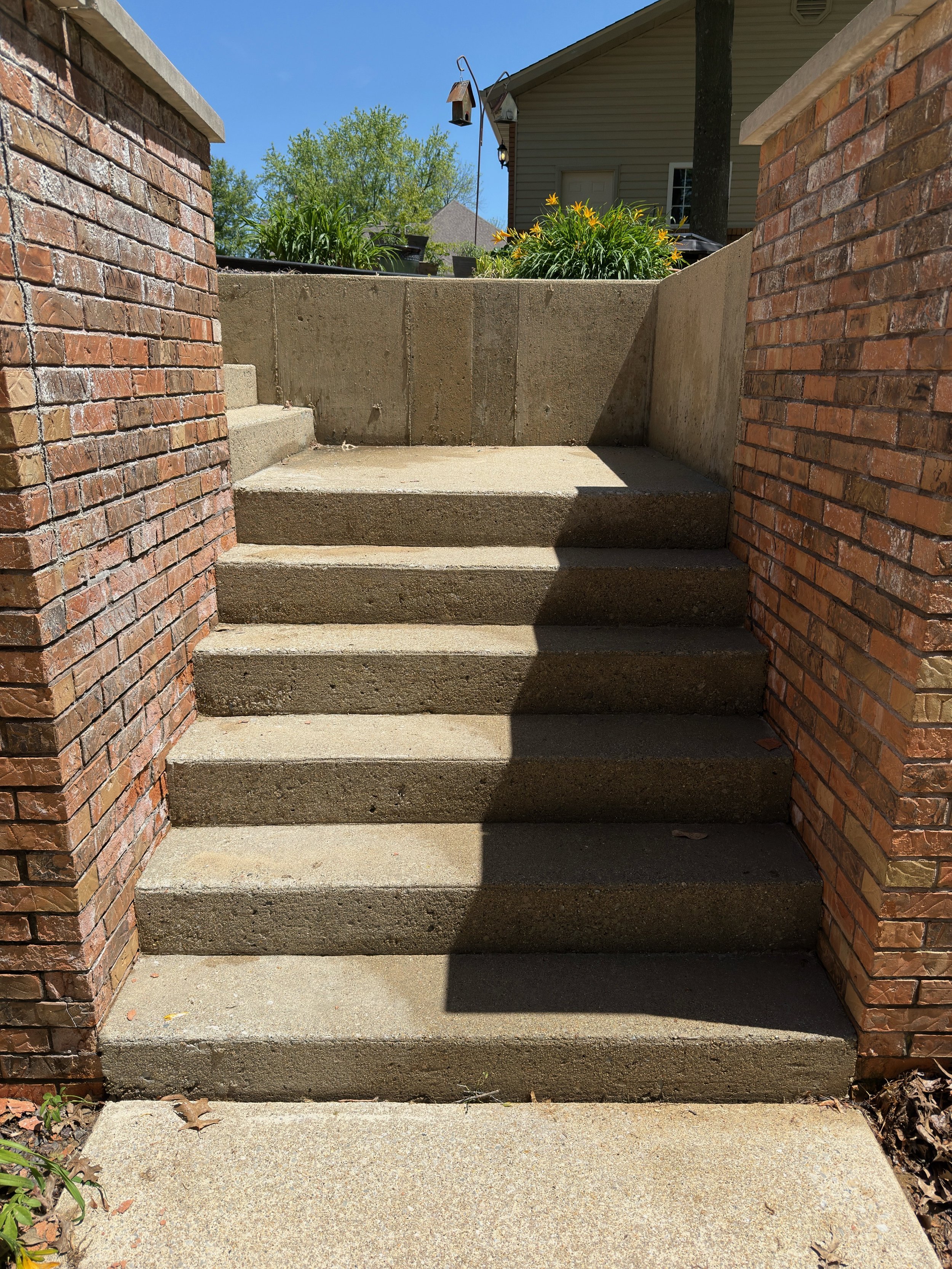 Concrete steps leading up between brick walls to a small landing with plants and a house in the background.