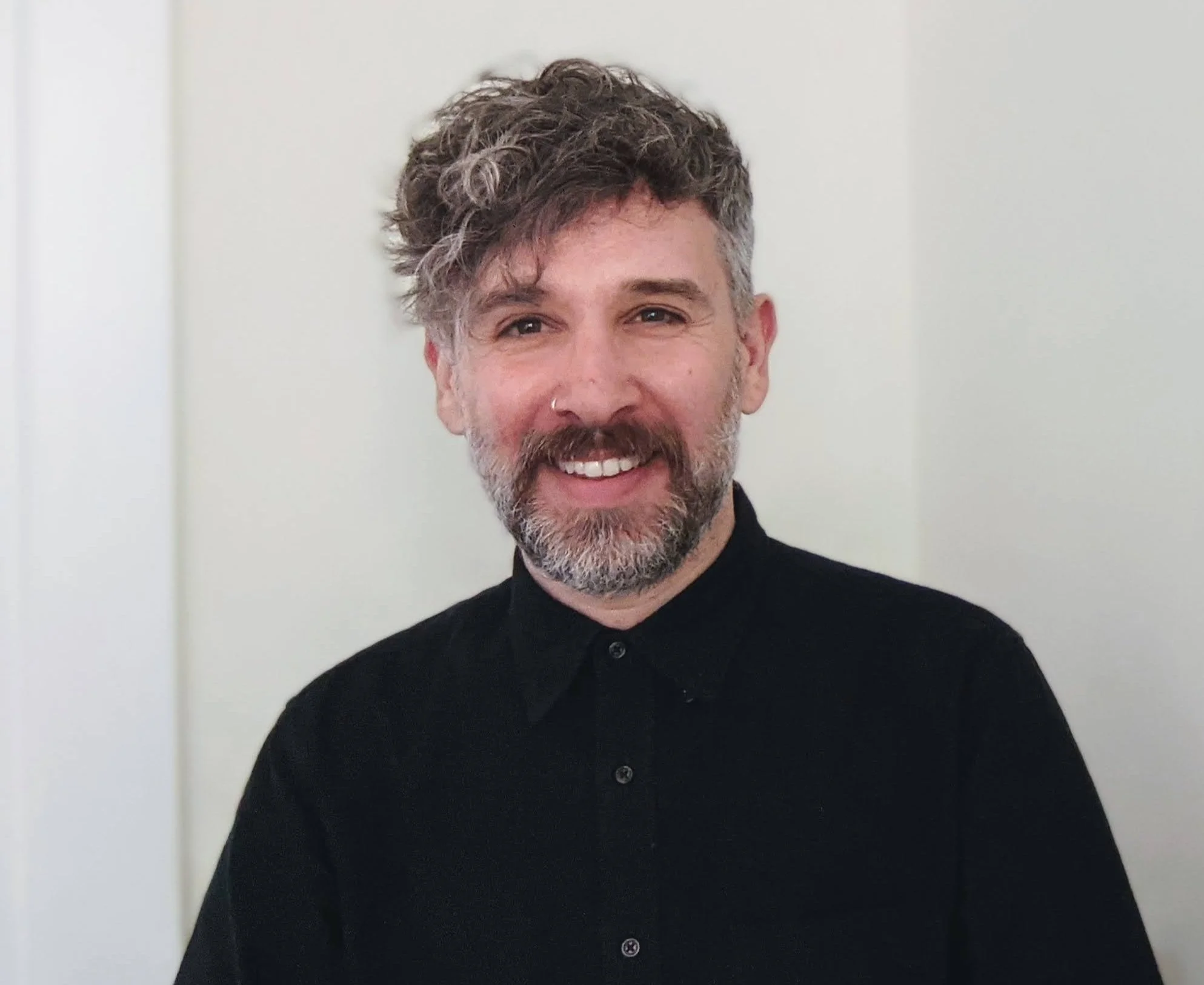 Smiling man with curly, graying hair and beard, wearing a black shirt, standing against a plain white background.