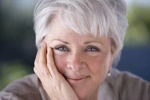 Close-up of a smiling elderly woman with short white hair, resting her face on her hand outdoors.