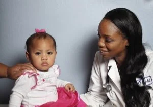 A female doctor or nurse with dark hair smiling at a young girl with pigtails and a pink bow, sitting on her lap in a medical setting.