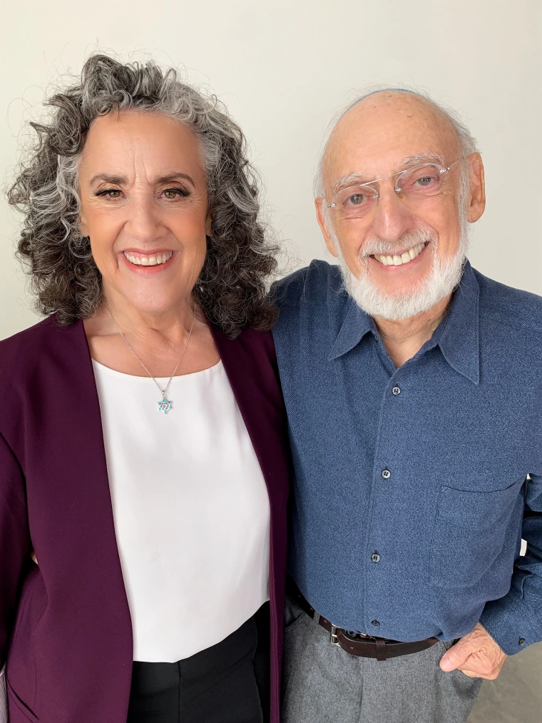 A smiling person with curly gray hair and another smiling person with glasses and a beard standing close together against a plain white background.
