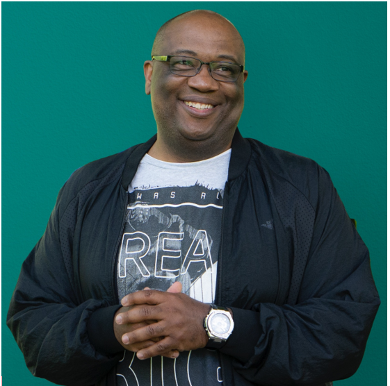 Smiling man wearing glasses, black jacket, white T-shirt, and watch against a teal background.