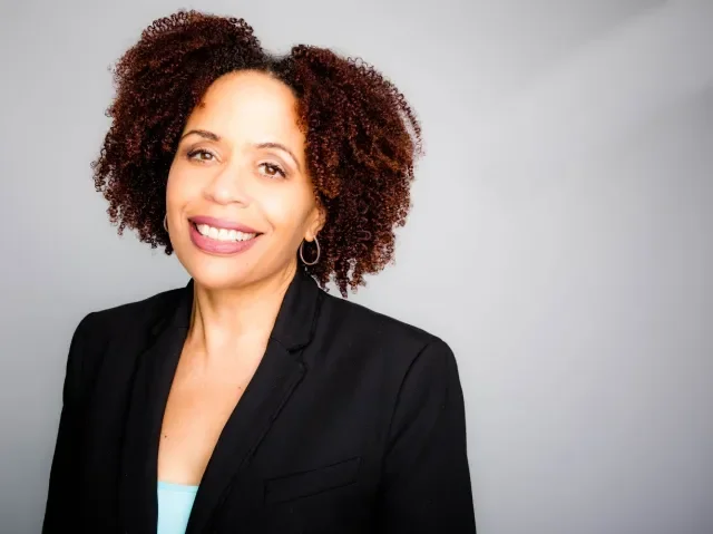 Portrait of a woman with curly hair, smiling, wearing a black blazer and a light blue top against a plain gray background.
