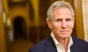 An older man with gray hair and a serious expression, wearing a suit jacket and white shirt, standing in a wood-paneled room.