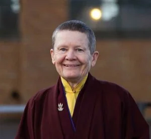 A woman with short gray hair wearing a dark maroon jacket over a yellow shirt, smiling outdoors.