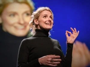 A woman with blonde hair speaking at a stage, with a large projection of her smiling face behind her, against a blue background.