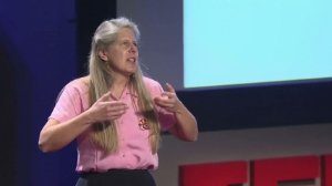 A woman with long blonde hair wearing a pink shirt speaking in front of a large screen during a presentation.