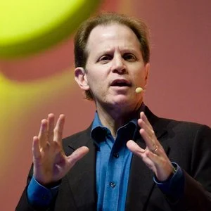 A man in a dark blazer and blue shirt speaking and gesturing with his hands.