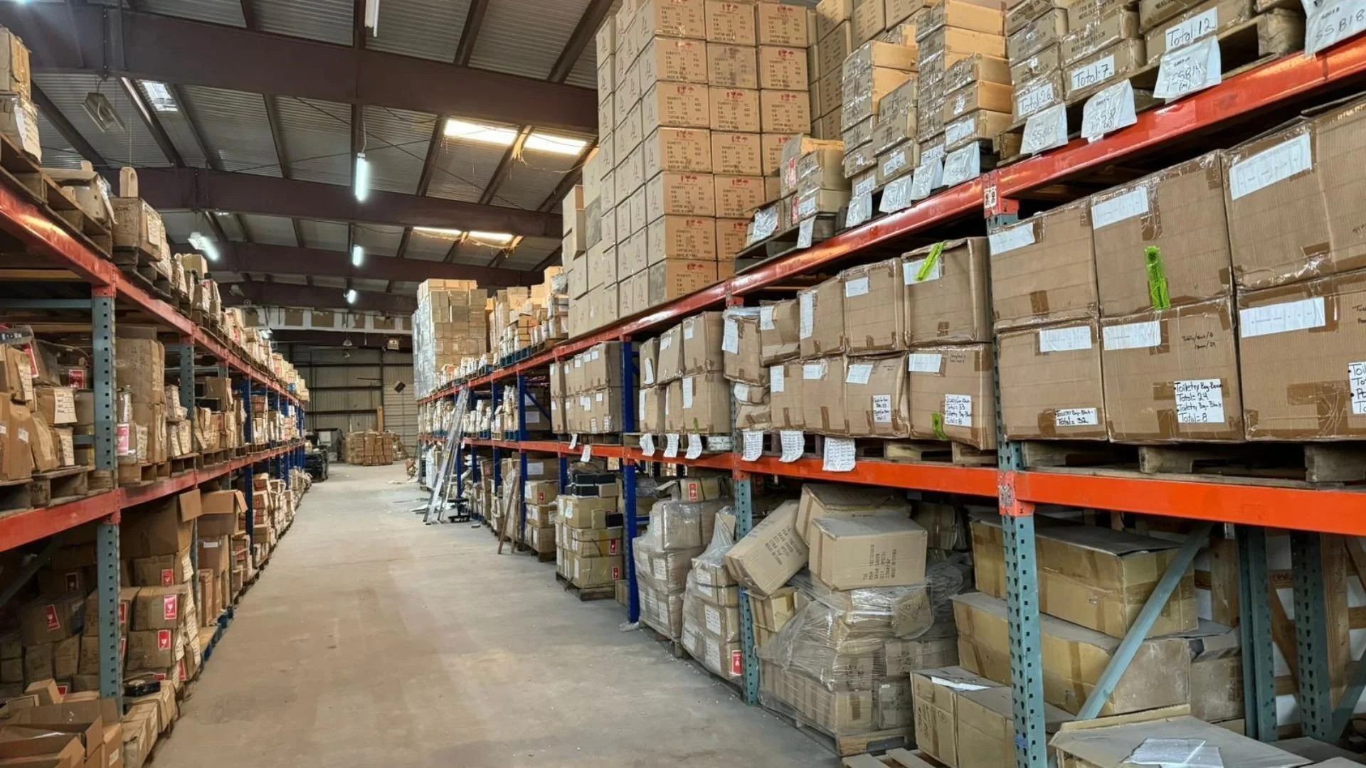 Warehouse aisle with metal shelving units filled with boxed and packaged products, some with labels and notes, extending into the distance under a high ceiling with fluorescent lighting.