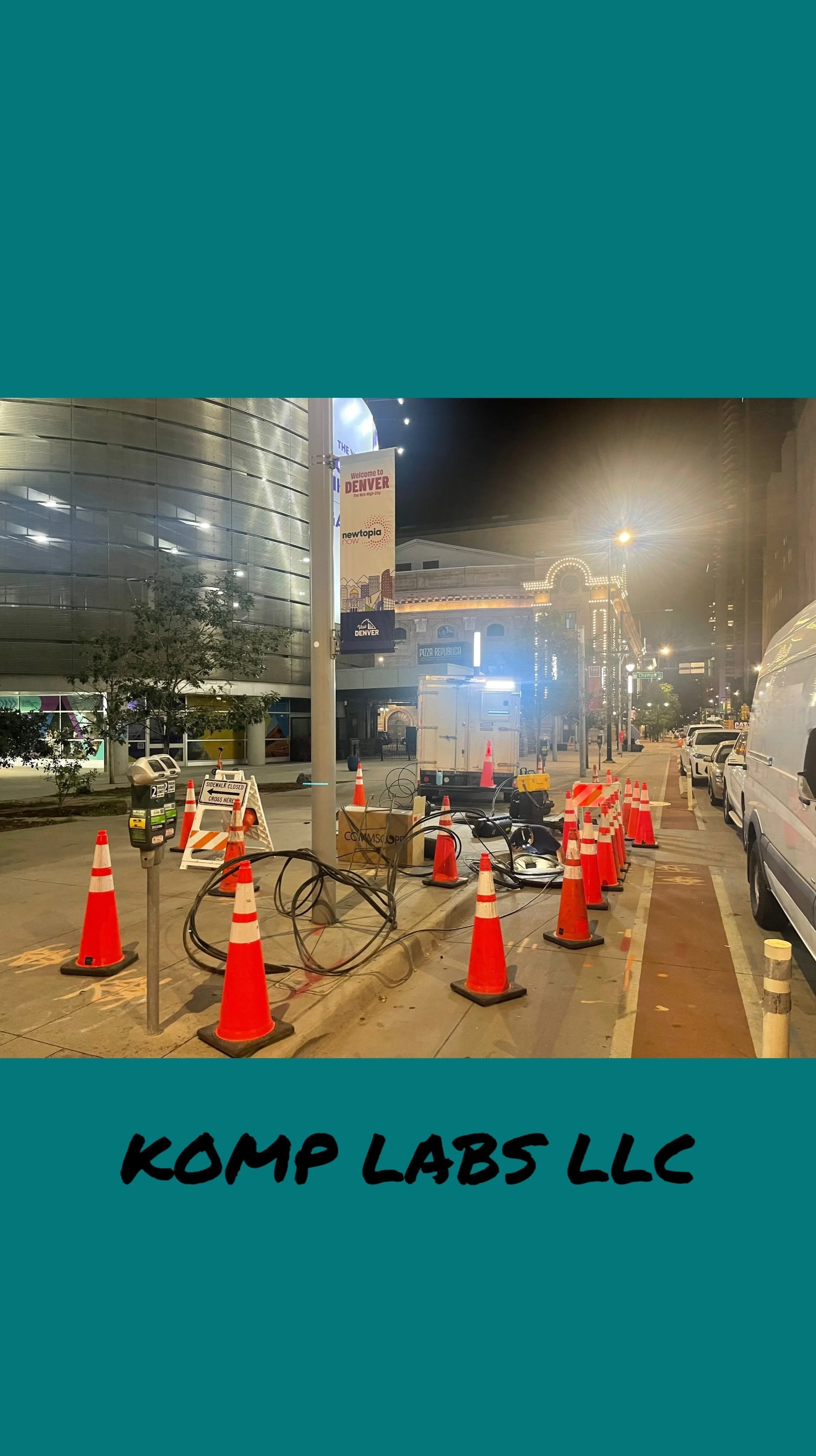 Night scene of construction or maintenance work on a city street, with orange cones, cables, and equipment, in front of modern buildings with bright signage and streetlights.