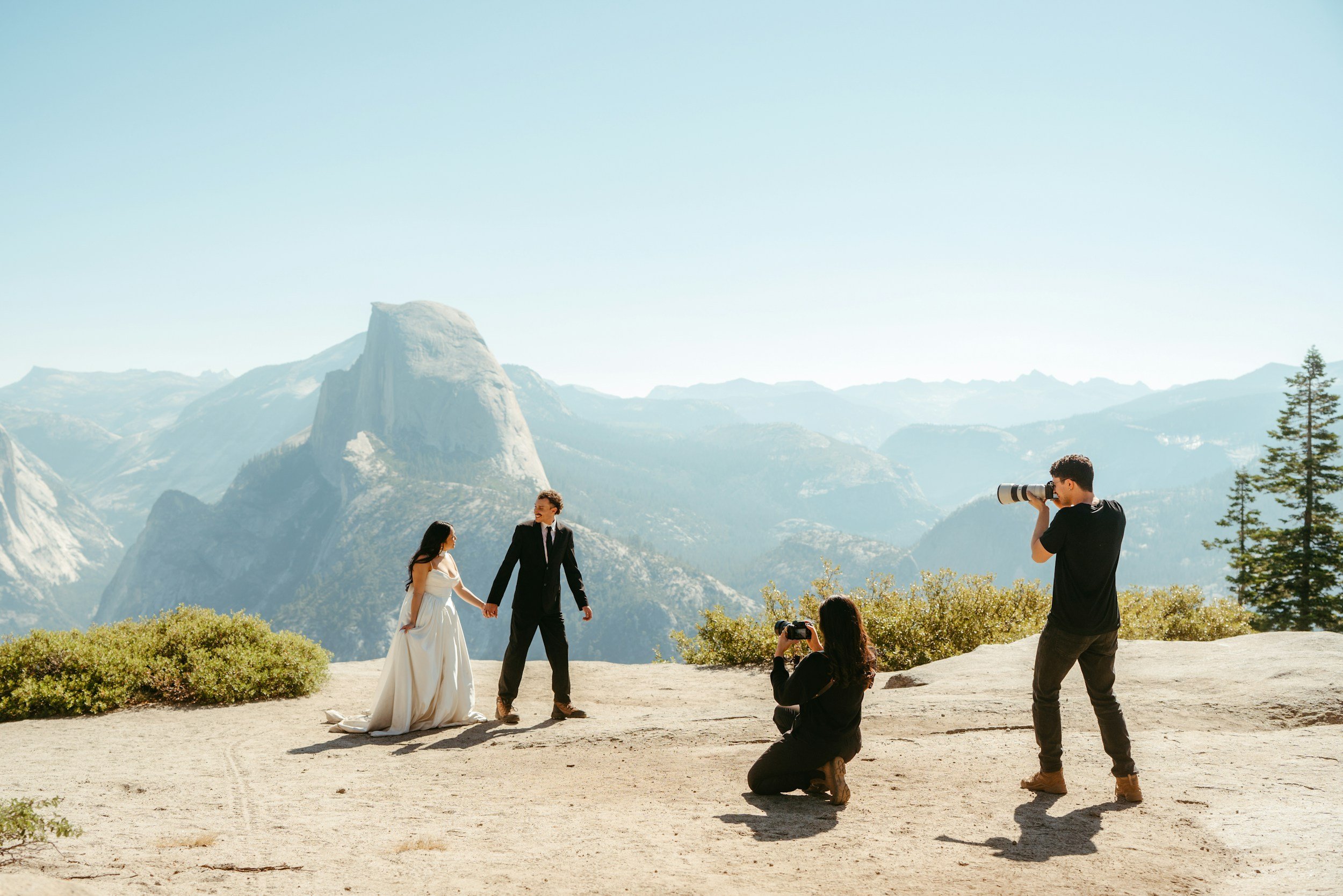 A wedding photoshoot happening outdoors in a mountainous area with two people dressed in wedding attire, holding hands, and posing for photographers with mountain scenery in the background.