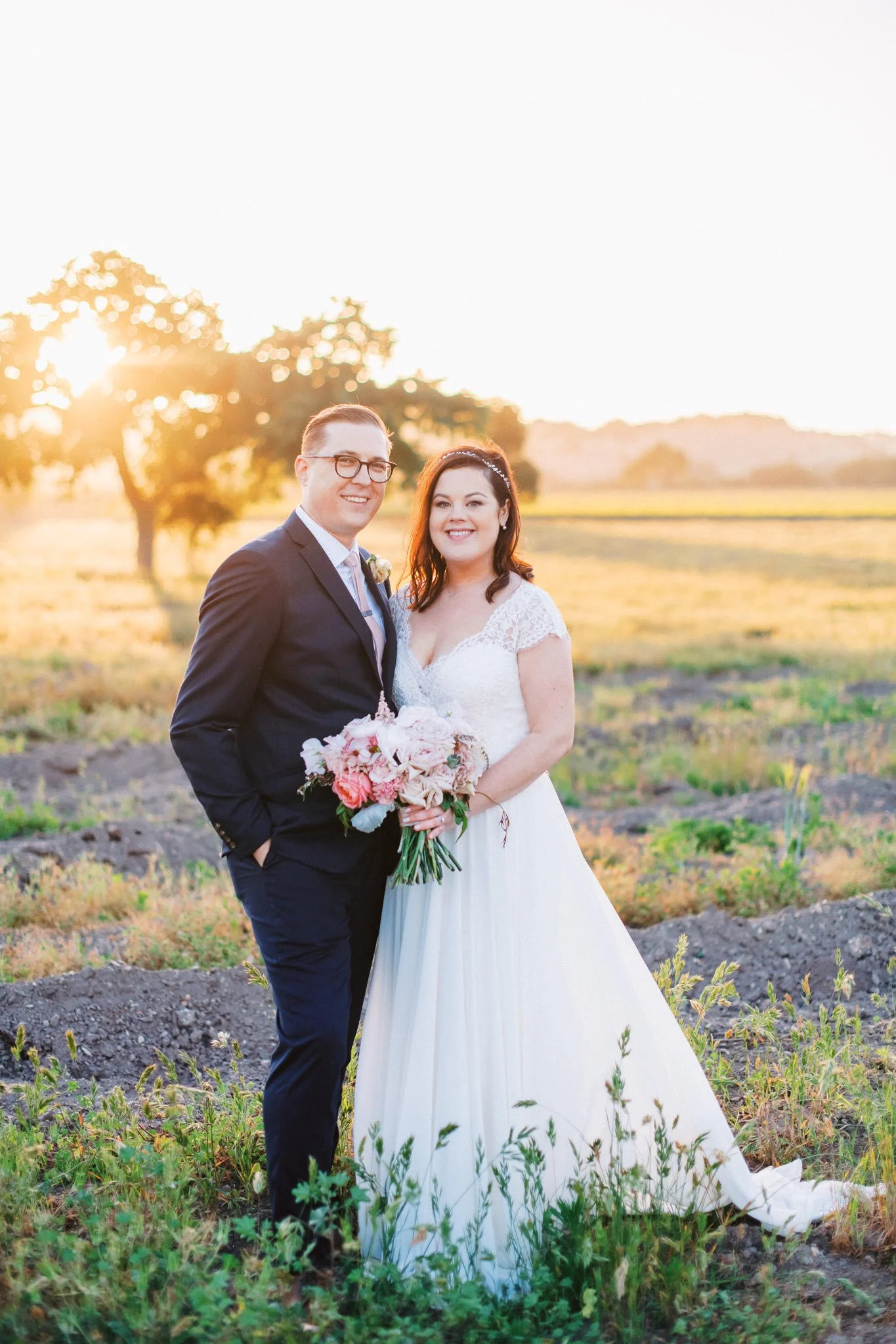 A newlywed couple stands outdoors during sunset, with the groom in a dark suit and glasses, and the bride in a white wedding dress holding a bouquet of pink and white flowers.