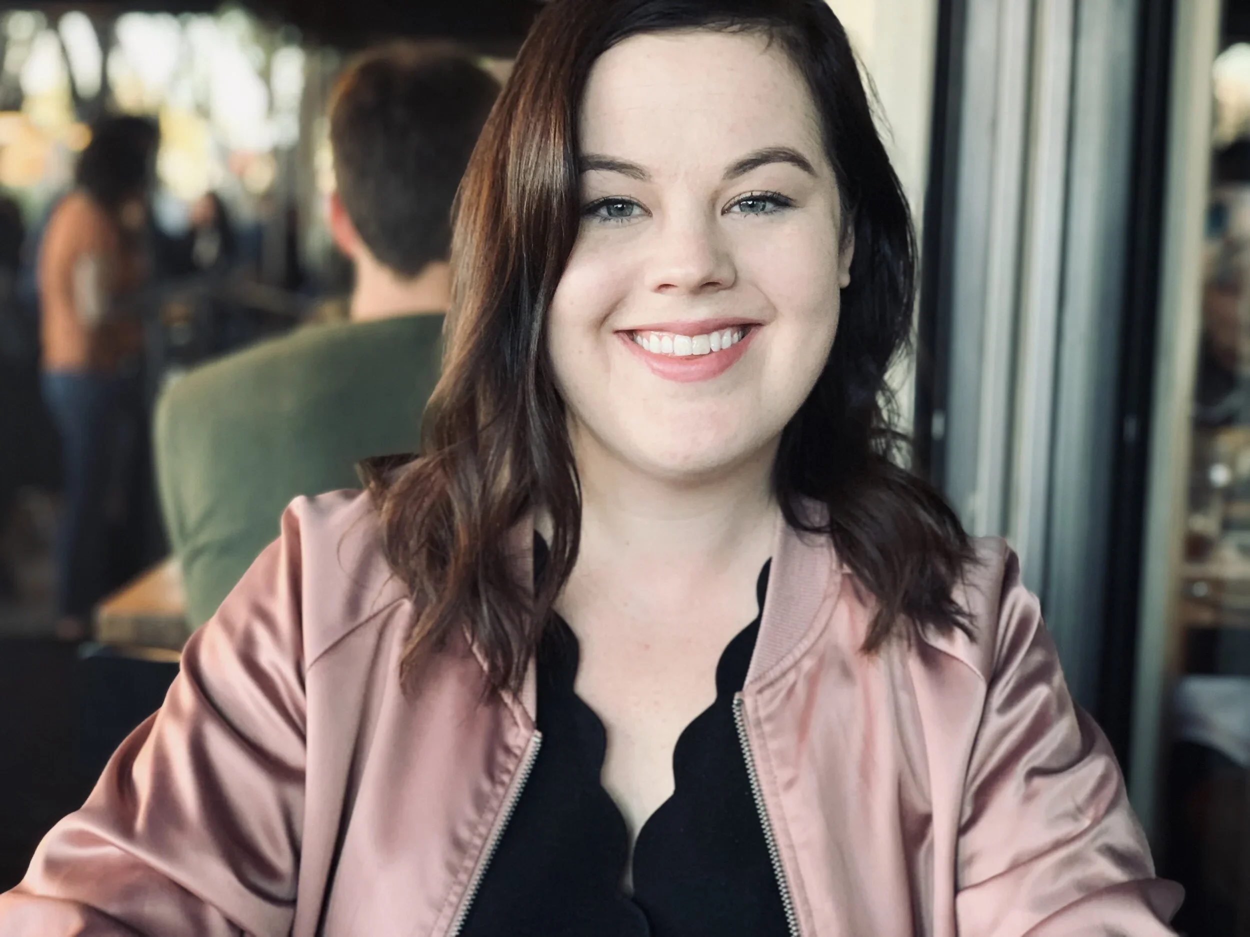 A woman with shoulder-length dark hair smiling in a restaurant or cafe setting, with blurred background of people and large windows.