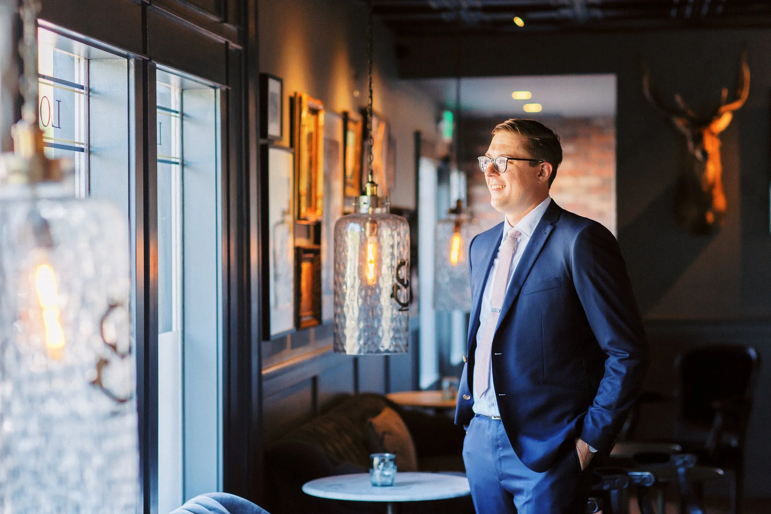 Man in a blue suit and glasses smiling while looking out a window in a stylish restaurant or cafe.