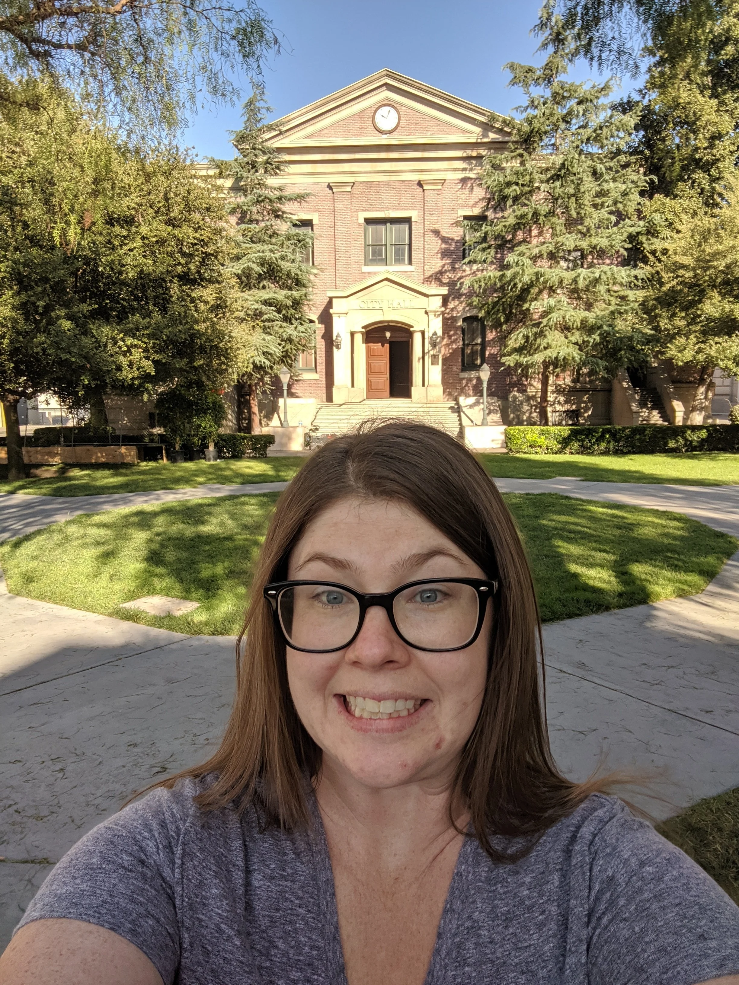 A woman with brown hair and glasses taking a selfie in front of the Back to the Future brick city hall building with trees and a green lawn on the Universal Studios Hollywood back lot.