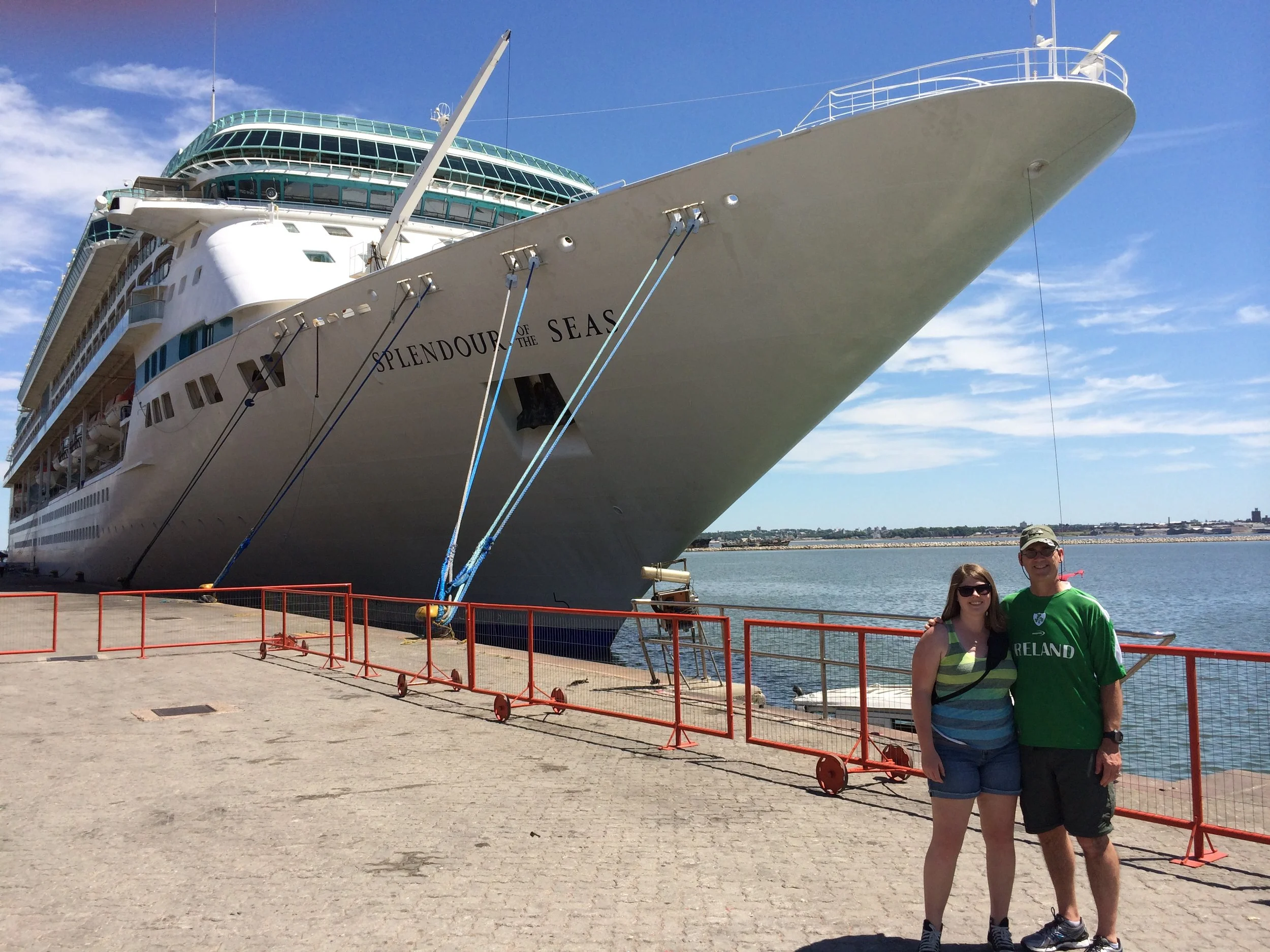 A large cruise ship named 'Splendour of the Seas' docked at a port with a woman and man standing in front of a red safety barrier, smiling and posing for a photo. The ship is white with multiple decks and a glass-enclosed area on top. The sky is clear with a few clouds and water is visible behind the couple.