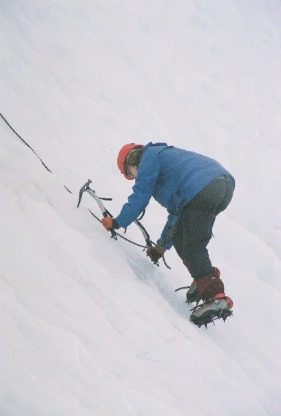 A woman using ice axe on a glacier slope while wearing crampons and a red helmet.