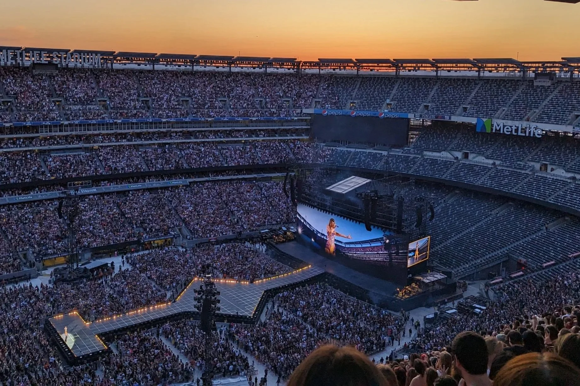 A large stadium filled with a crowd attending a concert at sunset, with a singer performing on a brightly lit stage with a big screen behind her.
