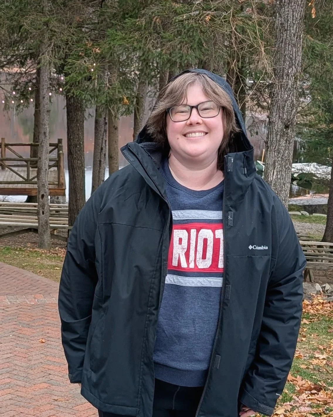 A woman with glasses and shoulder-length hair, smiling outdoors in a forested area, wearing a black jacket over a navy blue crewneck sweatshirt.