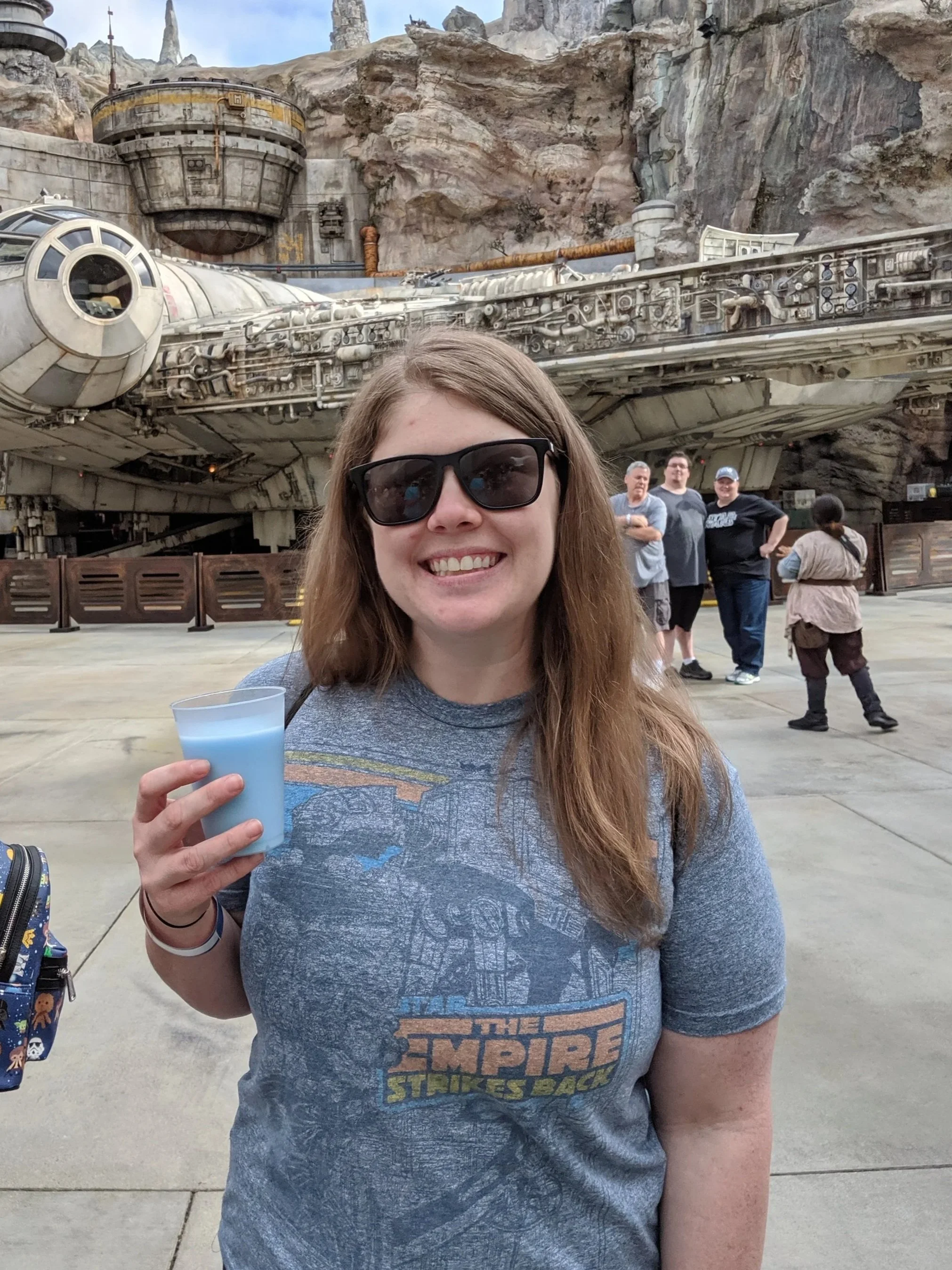 A woman with long brown hair, wearing sunglasses and a Star Wars t-shirt, smiling and holding a blue drink, standing in front of the Millennium Falcon at a theme park.