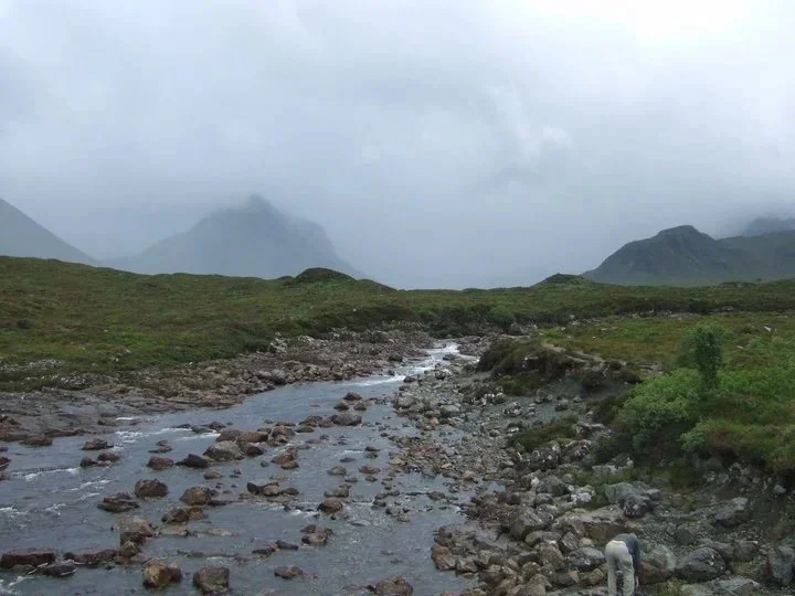 A mountain landscape with a rocky river flowing through green hills under a cloudy sky.
