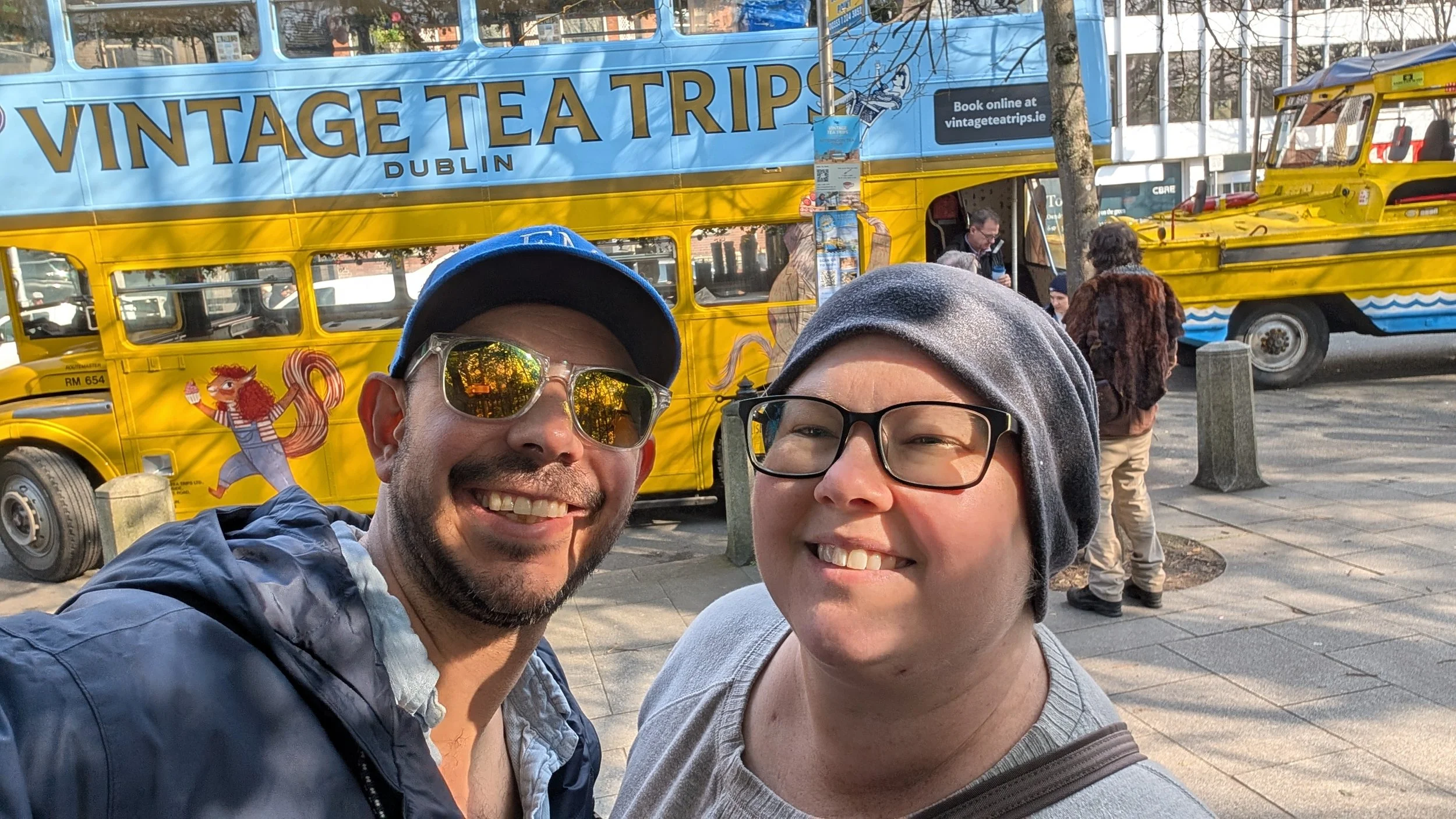 A couple taking a selfie on a city street. One wears a blue cap and sunglasses, the other a gray beanie and glasses. Behind them is a yellow vintage double-decker bus with the sign 'VINTAGE TEA TRIPS DUBLIN' and a cartoon squirrel illustration.