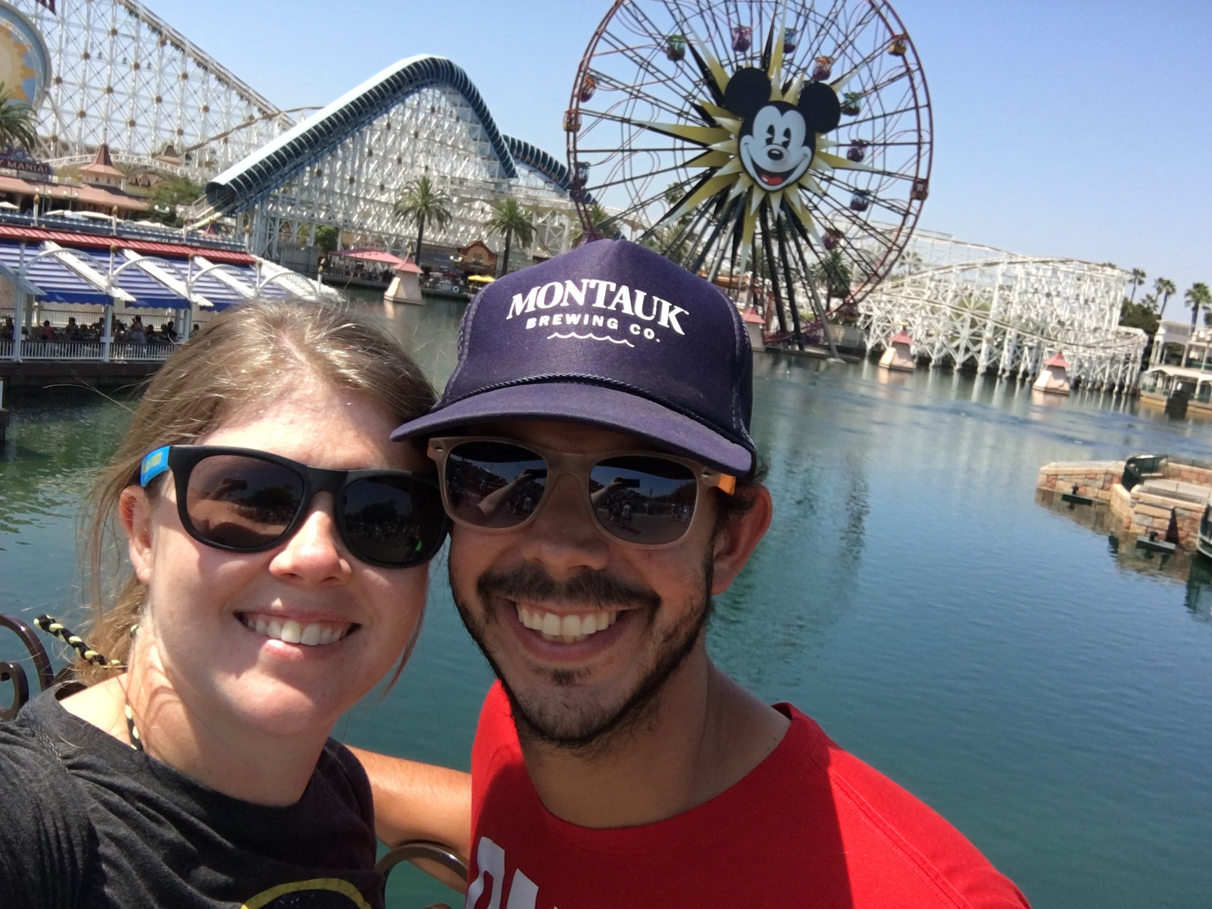 Smiling couple taking a selfie at an amusement park near a body of water, with a large Mickey Mouse Ferris wheel and roller coaster in the background on a sunny day.