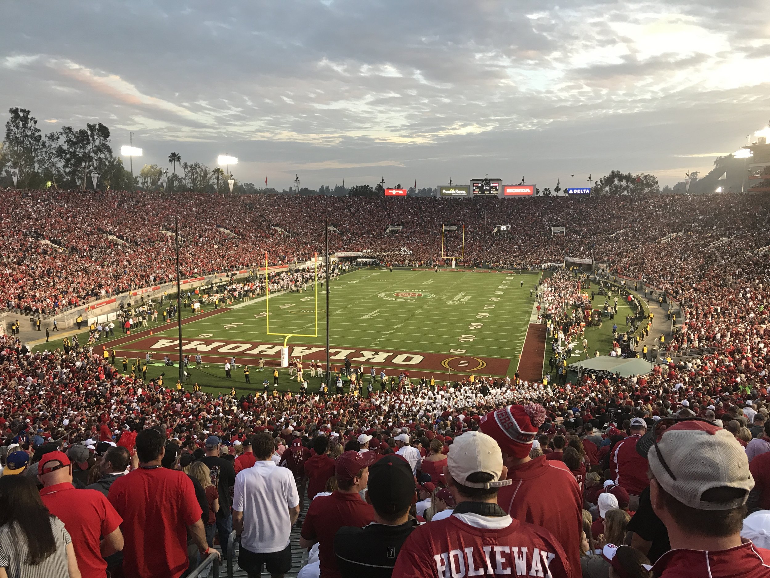 A crowded football stadium filled with fans, with a field in the center, some players on the sidelines, and a sunset sky above.