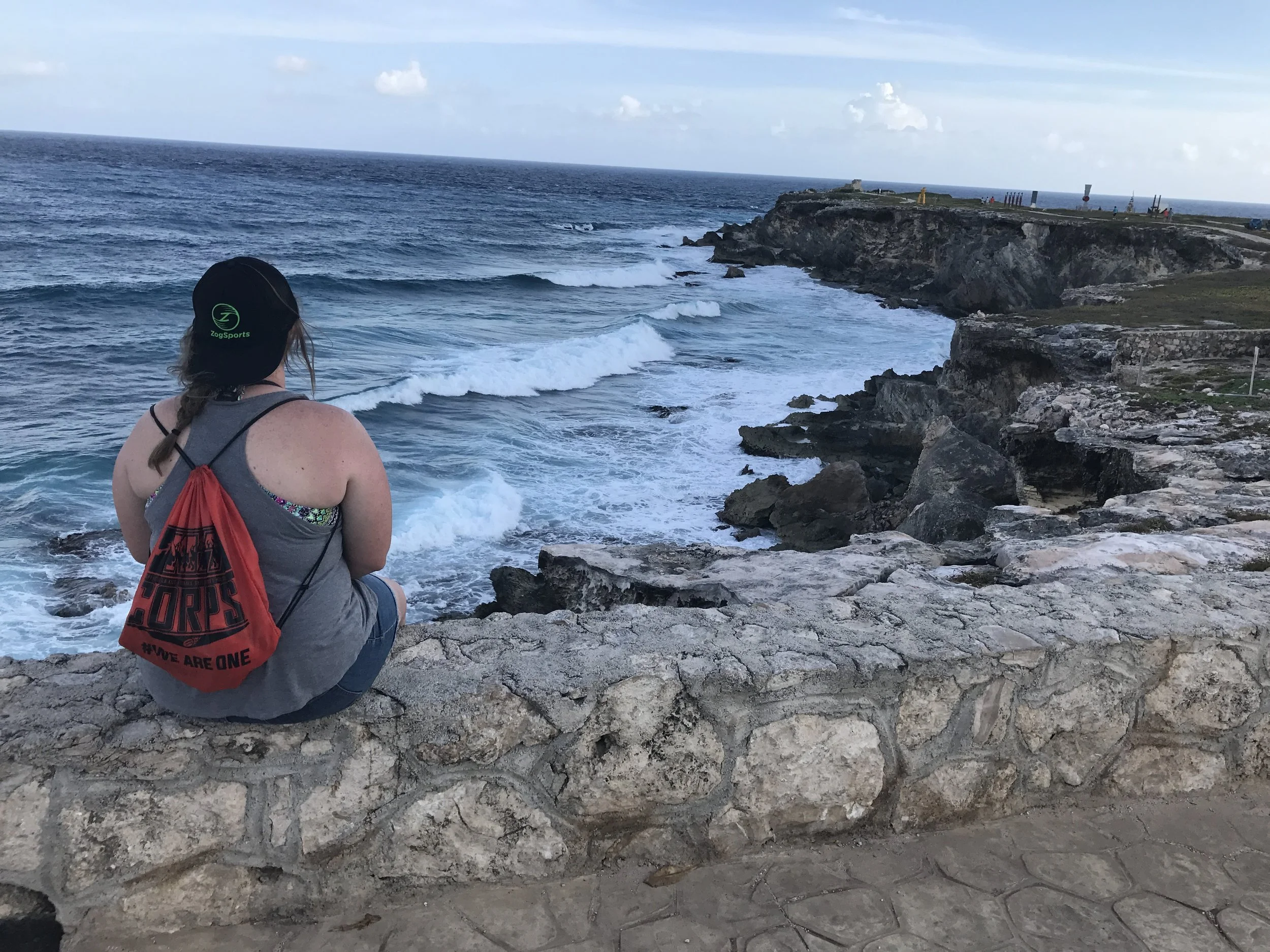A woman sitting on a stone wall overlooking the rocky coastline and ocean with waves crashing against the rocks. She is wearing a black hat, gray sleeveless top, and a red drawstring backpack.