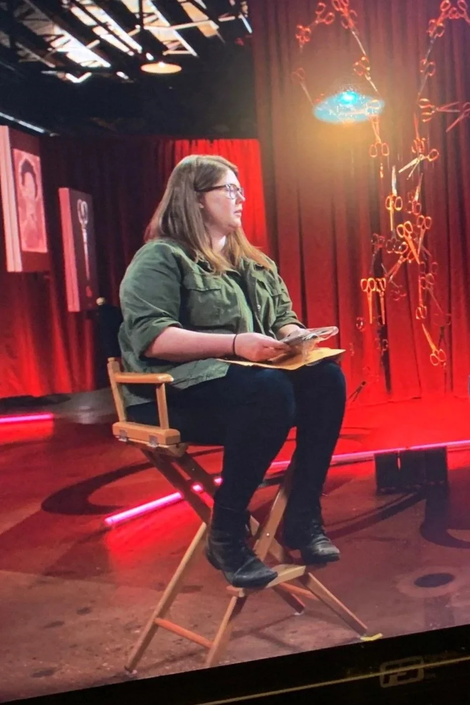 A woman with glasses, wearing a green jacket, sitting on a director's chair on a stage with red curtains and bright stage lighting, holding papers and a phone.