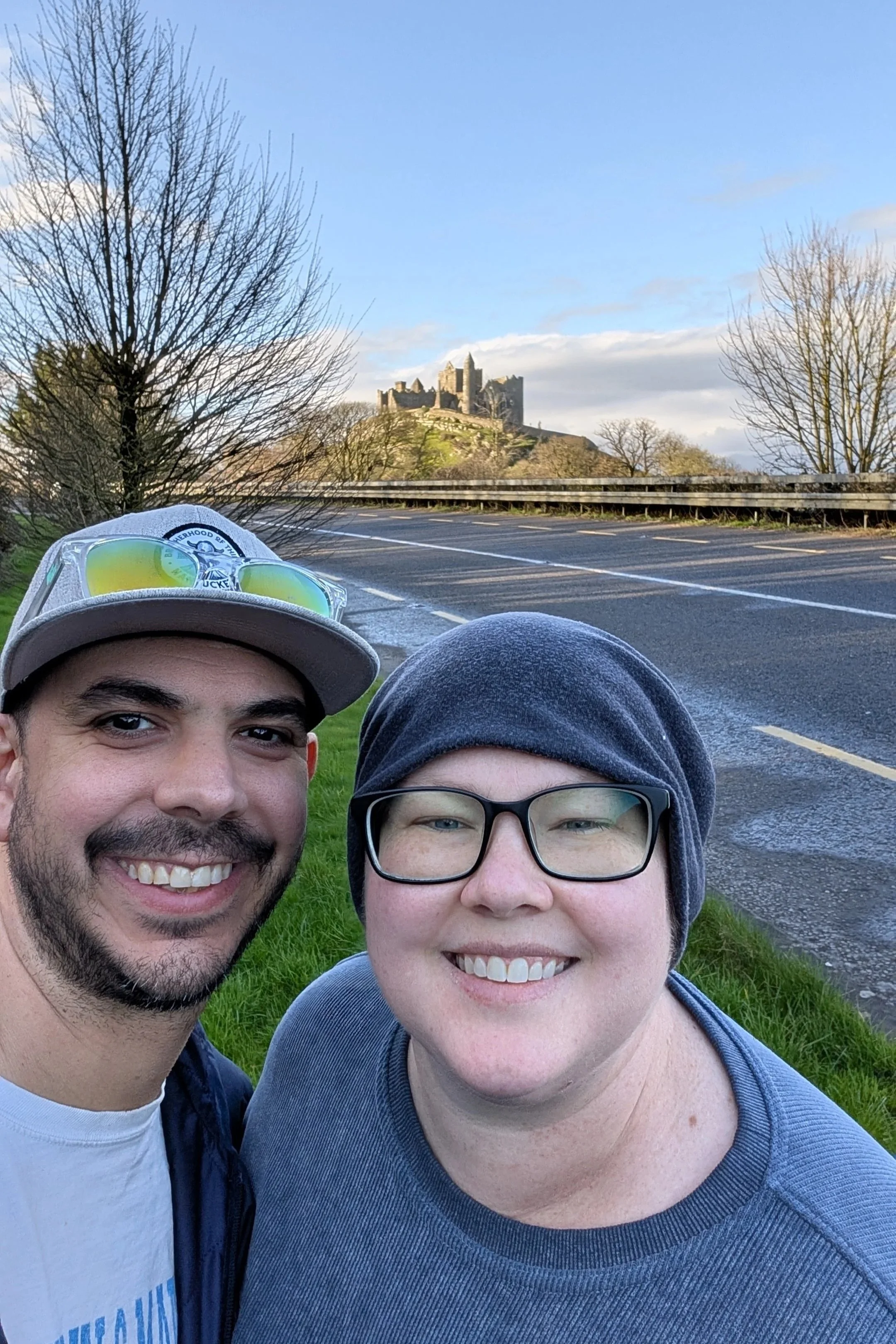 A couple taking a selfie outdoors with a castle on a hill in the background, leafless trees, and a paved road.
