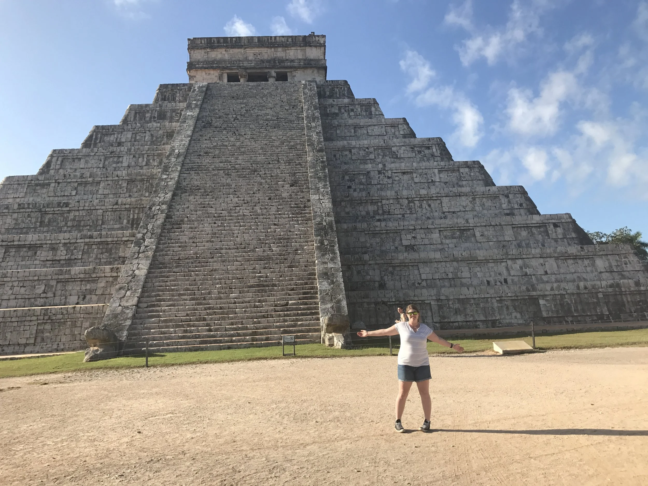 A woman standing with arms outstretched in front of the ancient Mayan pyramid at Chichen Itza, Mexico, under a blue sky with some clouds.