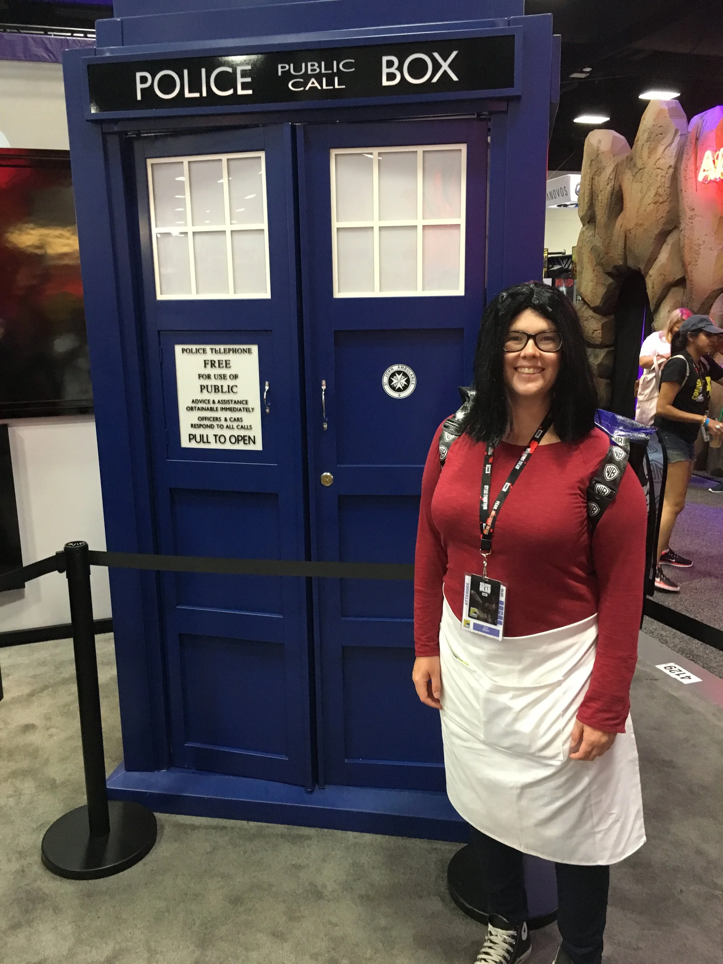 A woman dressed as a Linda from Bob's Burgers standing in front of a blue TARDIS police box at San Diego Comic-Con.