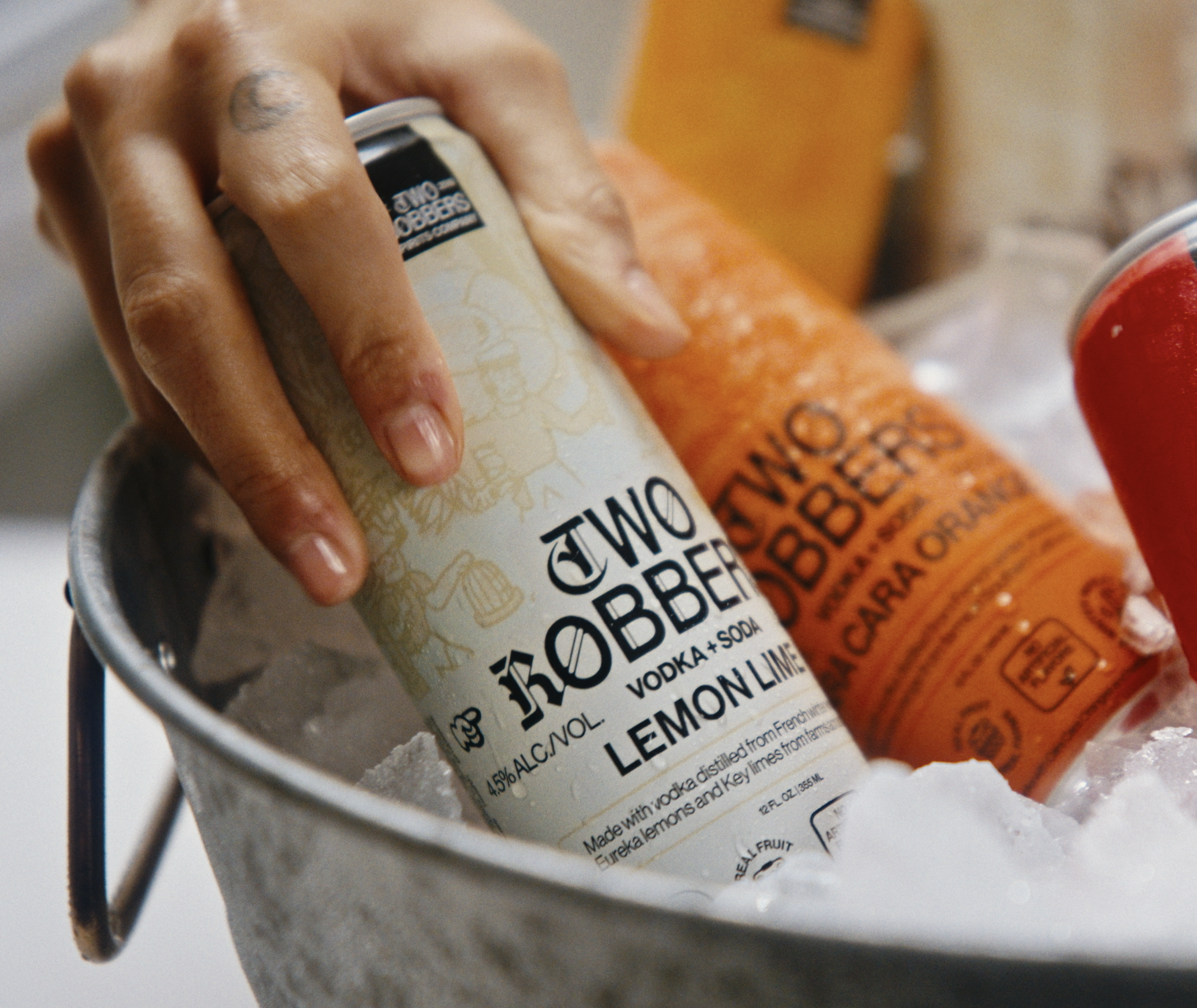 Person reaching into a refrigerator door, holding a box of colorful canned beverages labeled 'Two Robbers,' with lemons, limes, oranges, and a jar of pickles visible inside the fridge.
