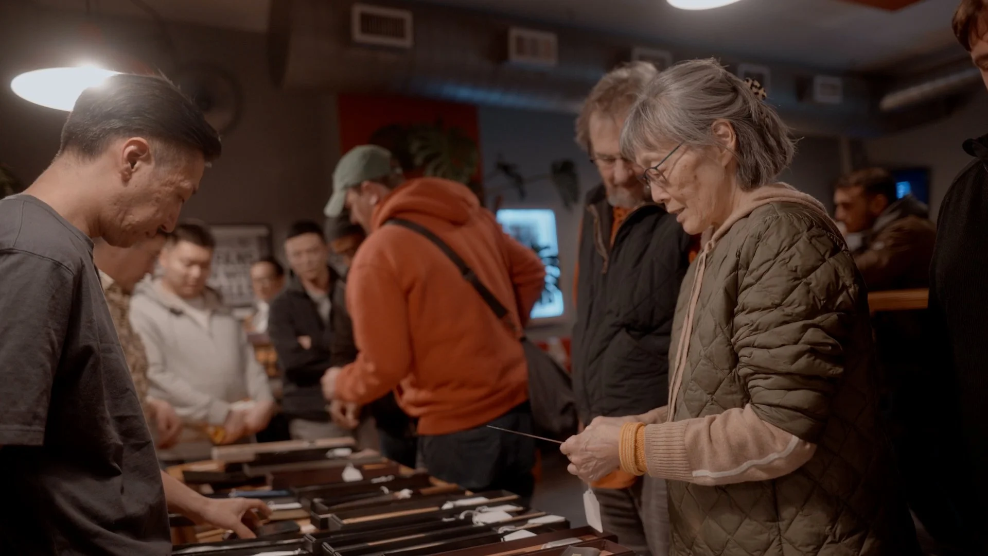 People browsing and examining knives or similar objects at a market or exhibit, with several individuals looking at items on display.