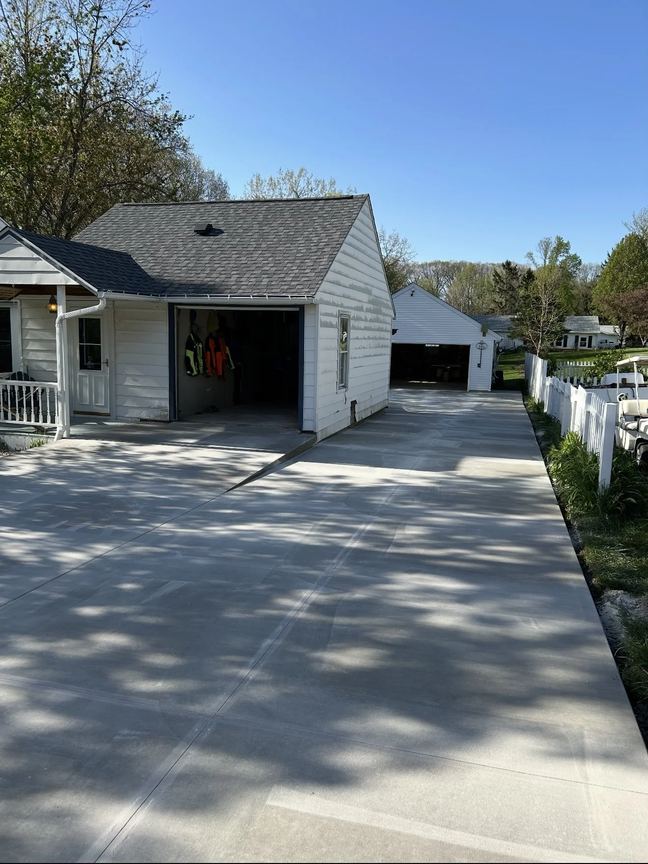 A house with a detached garage has a newly paved concrete driveway, shadowed by trees, with a white picket fence on the right side.