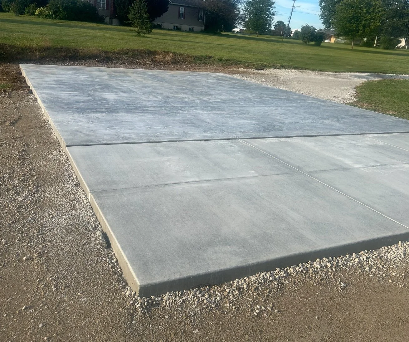 Freshly poured concrete slab in a grassy outdoor area with a house and trees in the background.