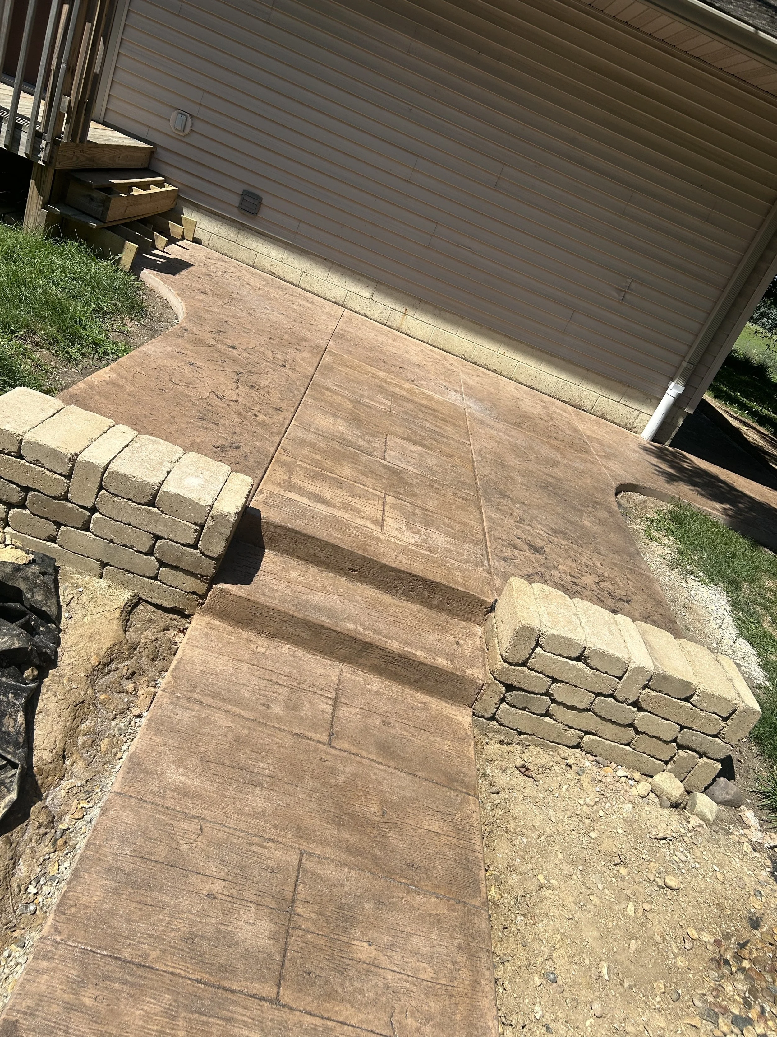 Newly constructed small concrete porch with steps leading up to a house, supported by brick walls on either side, with a concrete sidewalk extending from it.