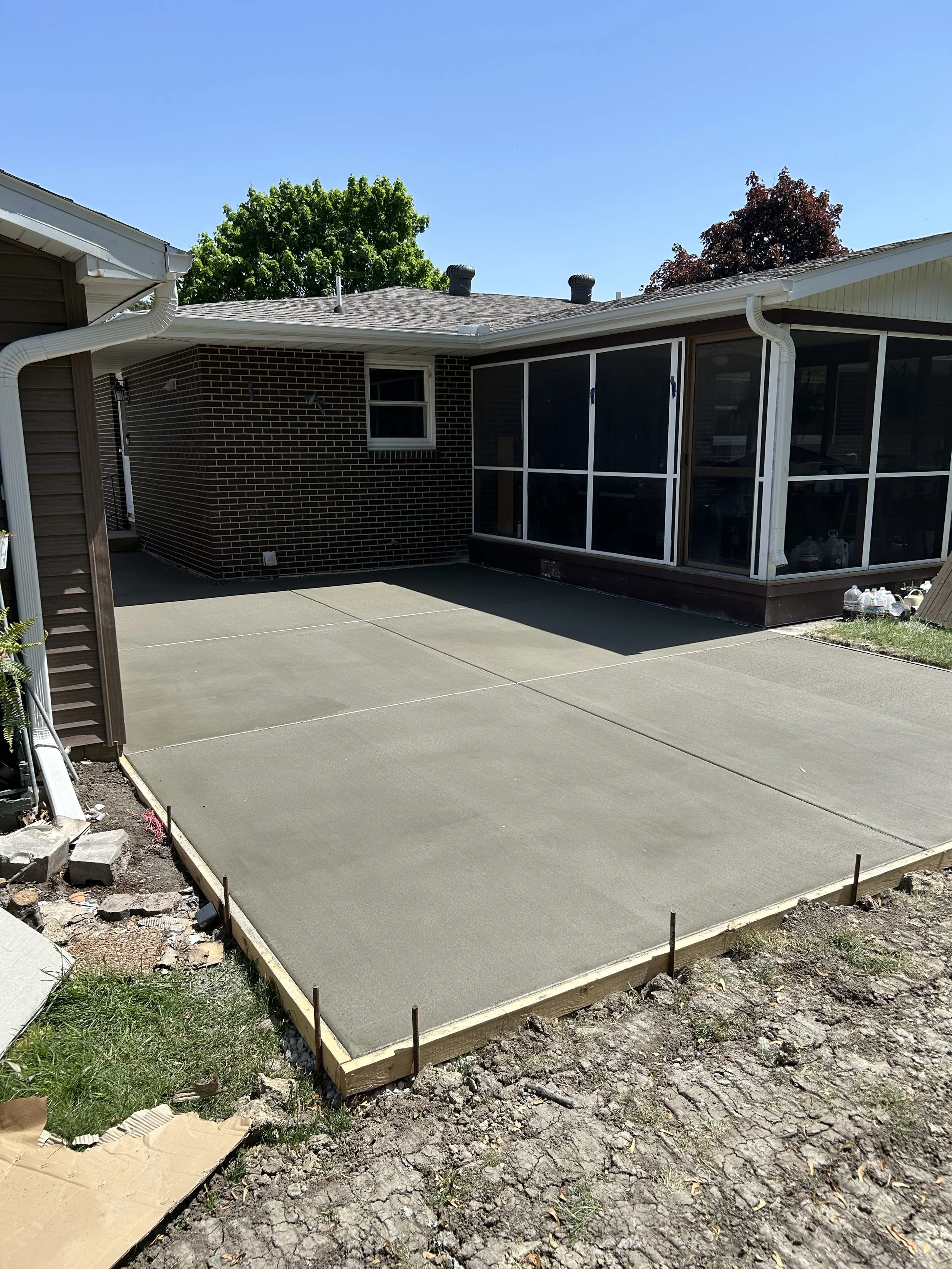 Freshly poured concrete patio with wooden frame in backyard of brick house, with screened porch and trees in the background.