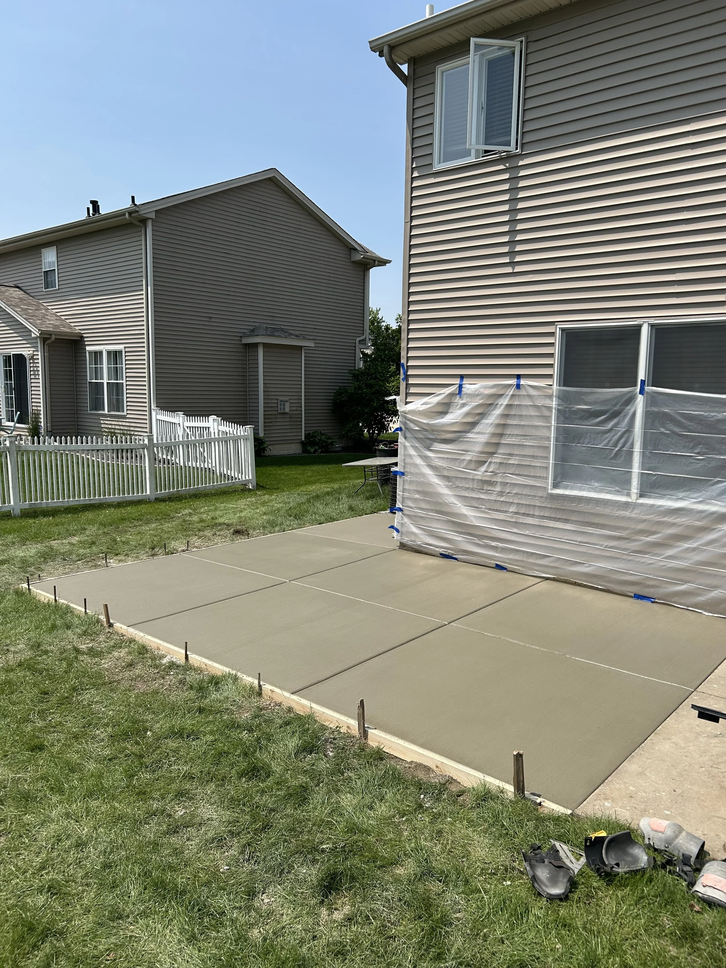 Freshly poured concrete patio in a backyard with a house in the background, surrounded by grass and neighboring homes, with construction shoes and tools on the grass.