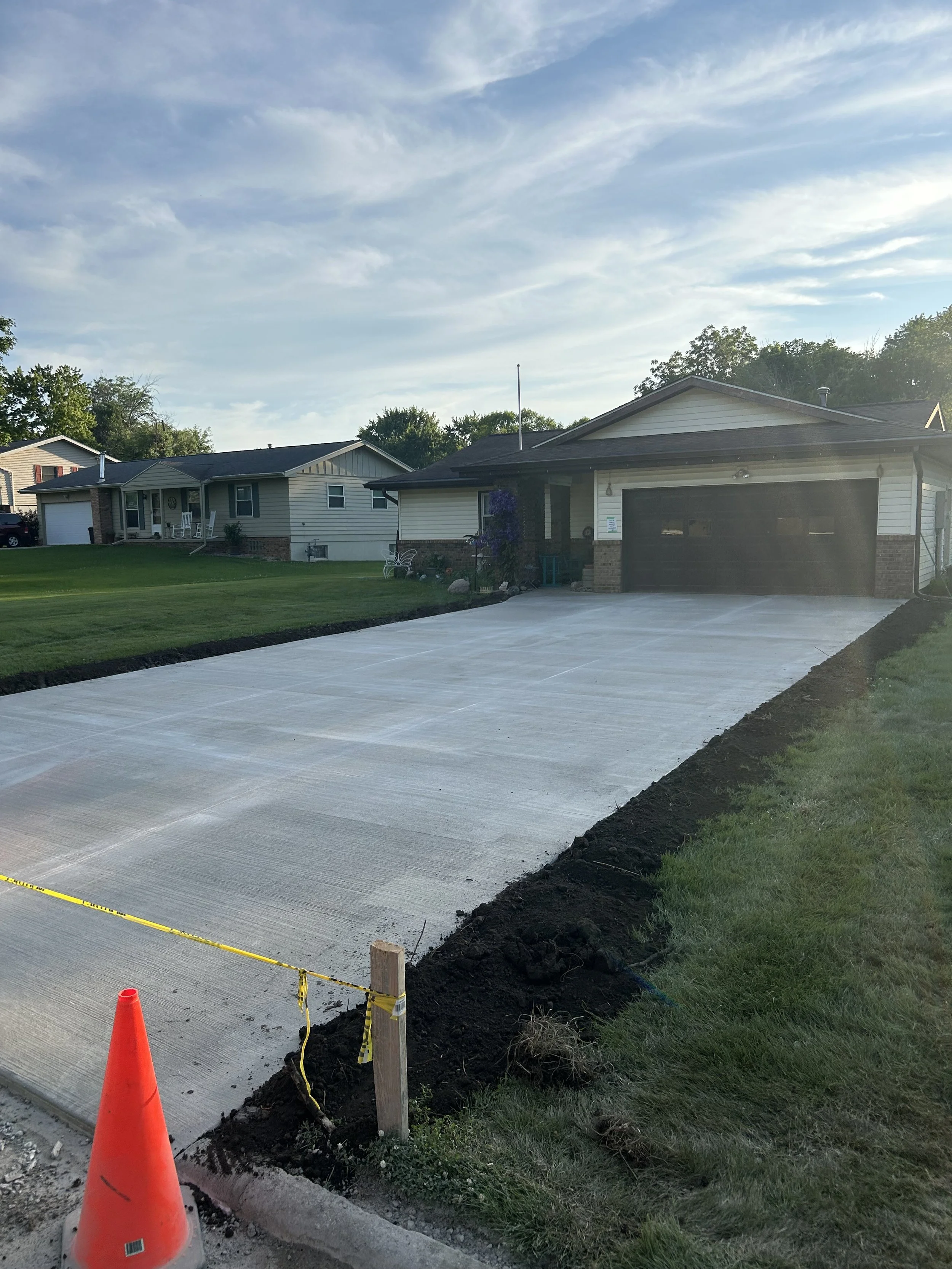 Newly poured concrete driveway in front of a suburban house under a partly cloudy sky, with construction cone and caution tape at the edge.