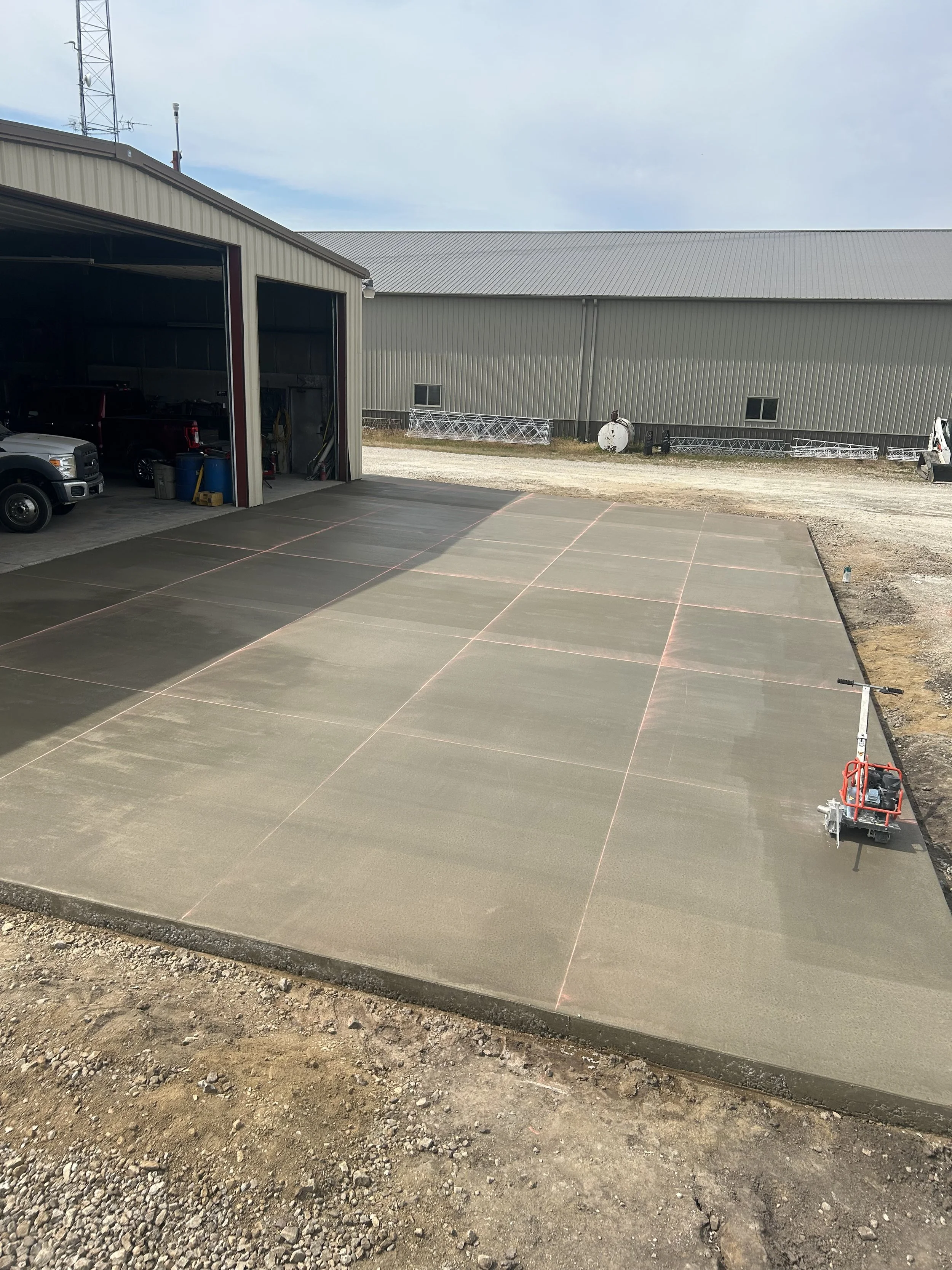 Newly poured concrete slab with red chalk lines marking sections outside a building, with construction equipment on the right and vehicles inside the building.