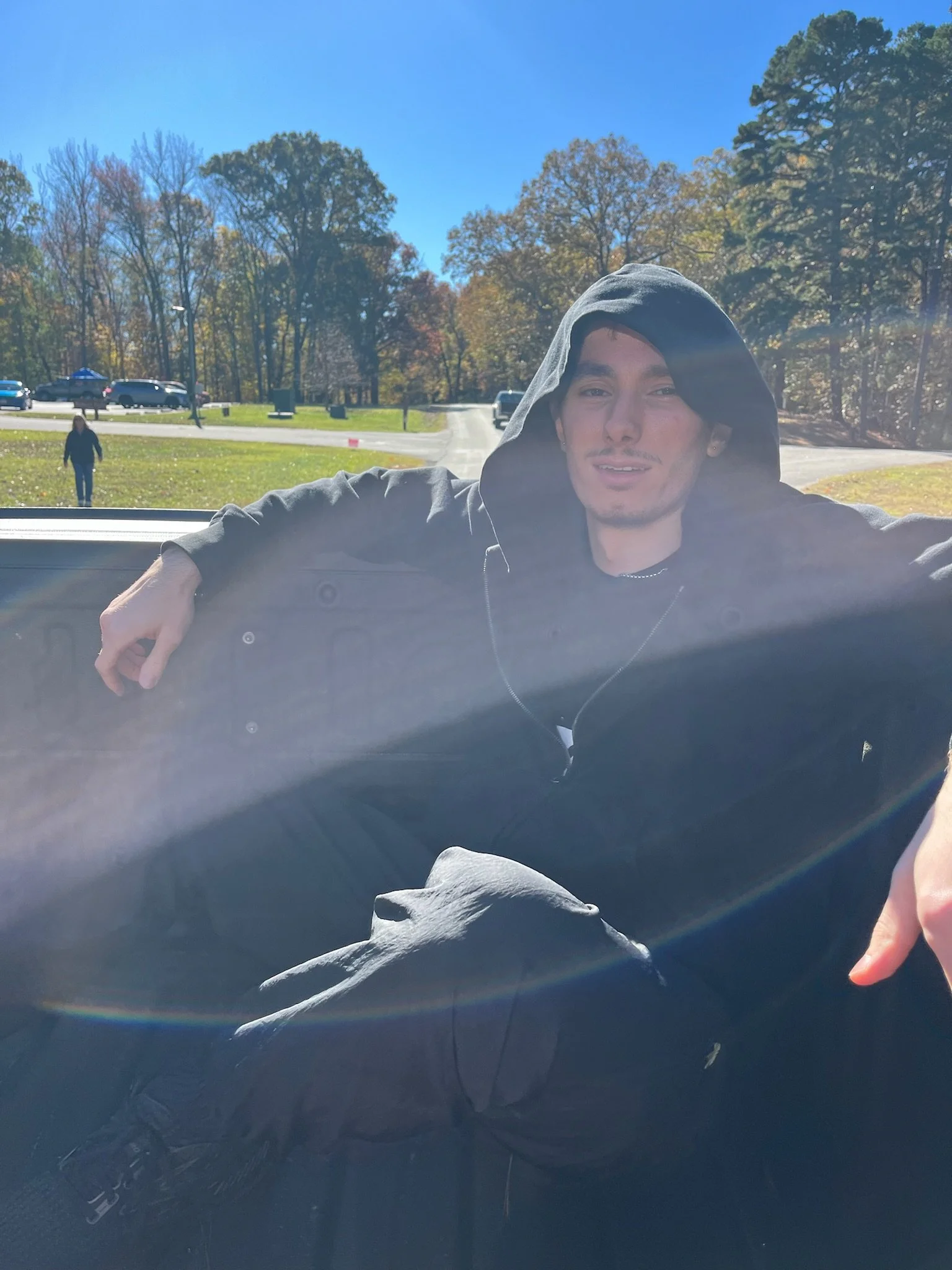 Young man in black hoodie and earrings sitting in the back of a vehicle on a sunny day with trees and parked cars in the background.