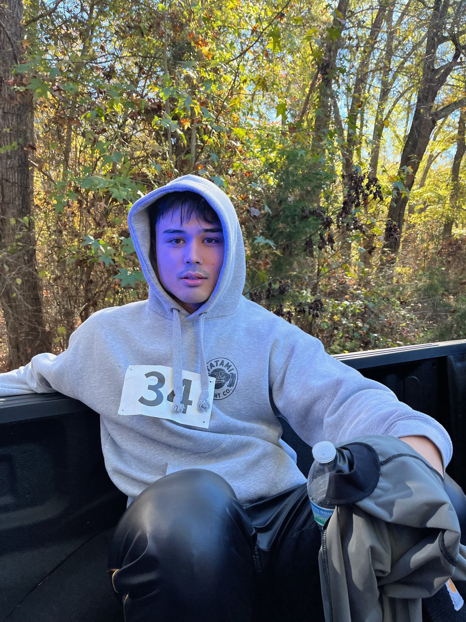 A young man wearing a gray hoodie with a race bib number 34, sitting in the back of a vehicle with a forest background, holding a water bottle and looking at the camera.