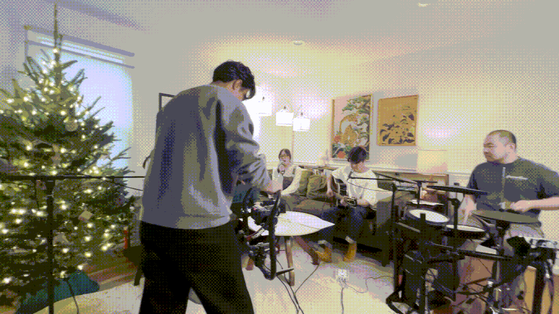 A person operating camera equipment in a living room decorated for Christmas, with a Christmas tree and four people sitting on a couch and chairs.