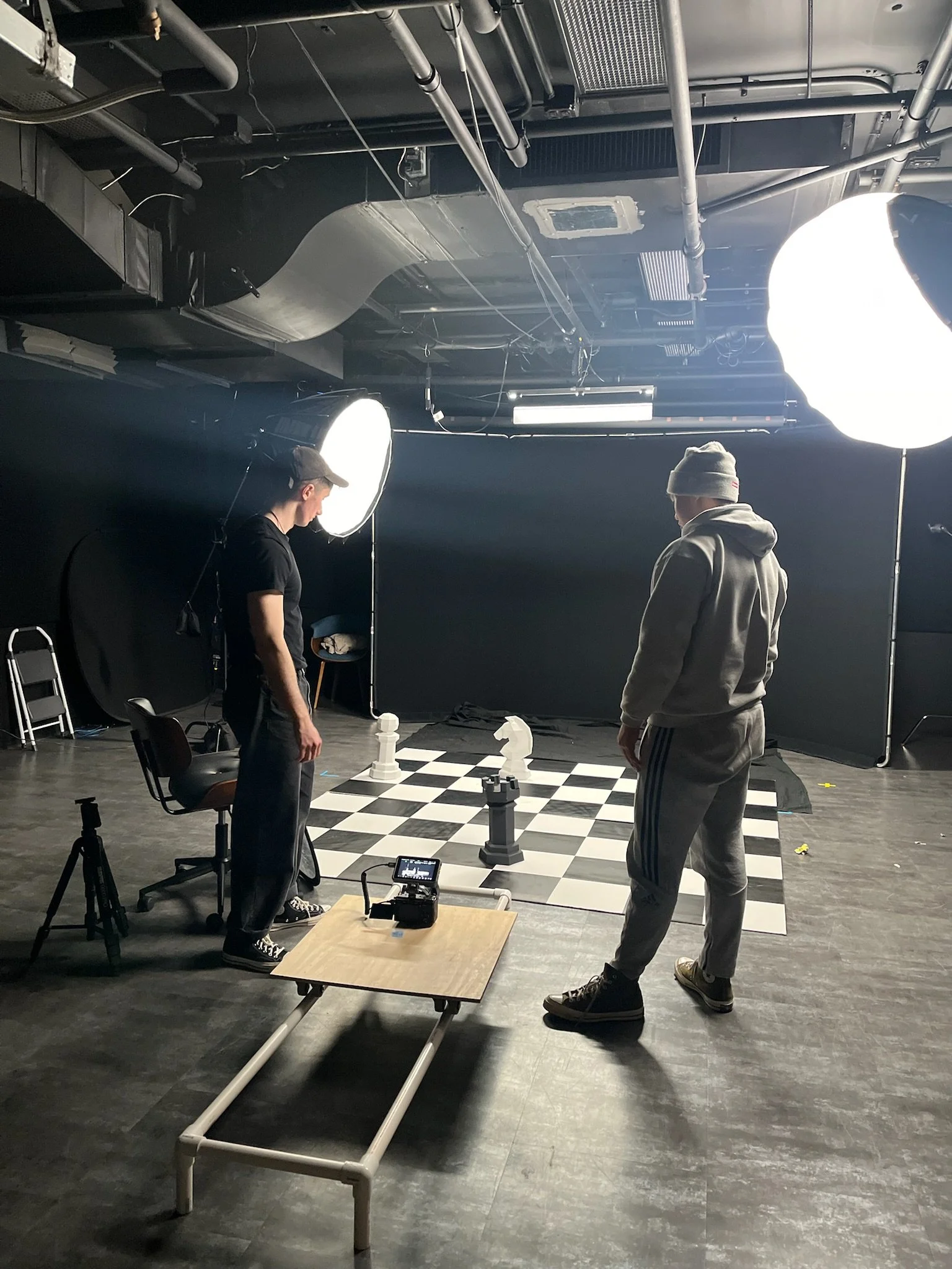 Two men standing on a chessboard set up for a chess game, with large studio lights, cameras, and equipment surrounding them in a photo studio.