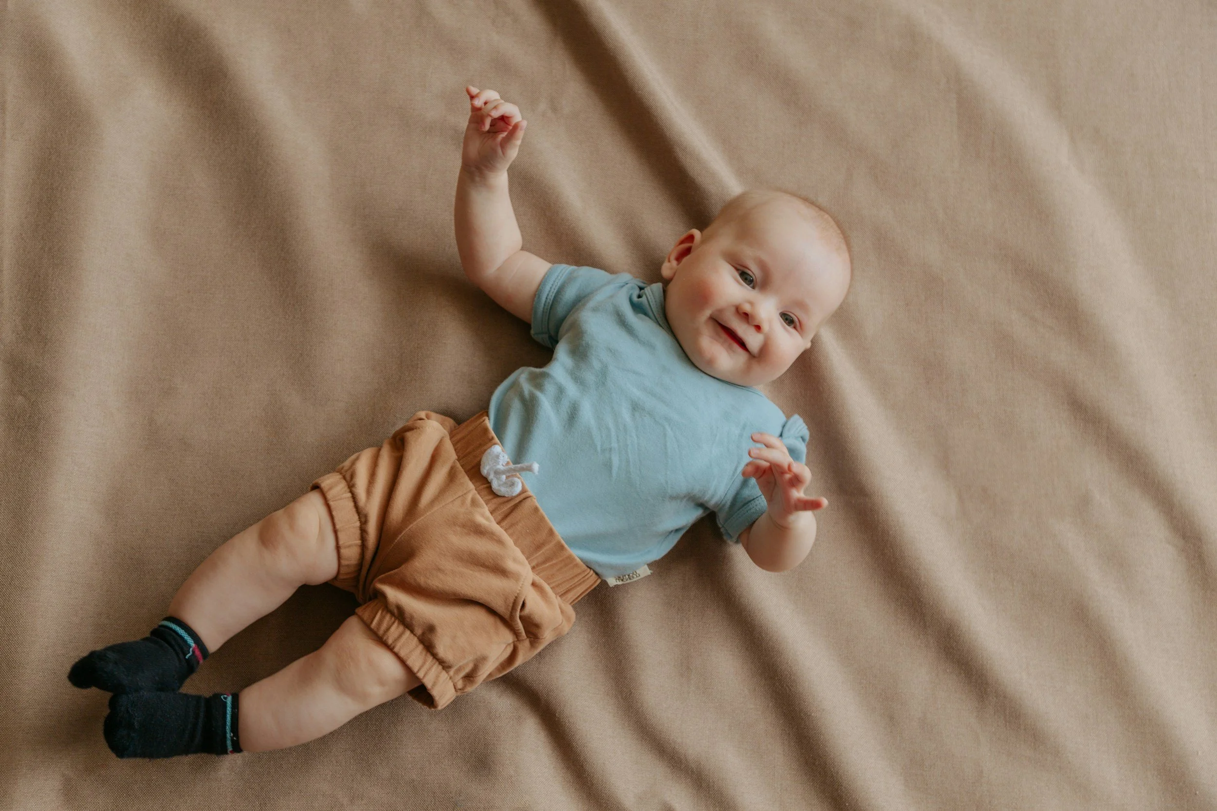 A smiling baby lying on a beige blanket, wearing a light blue shirt, brown shorts, and black socks.