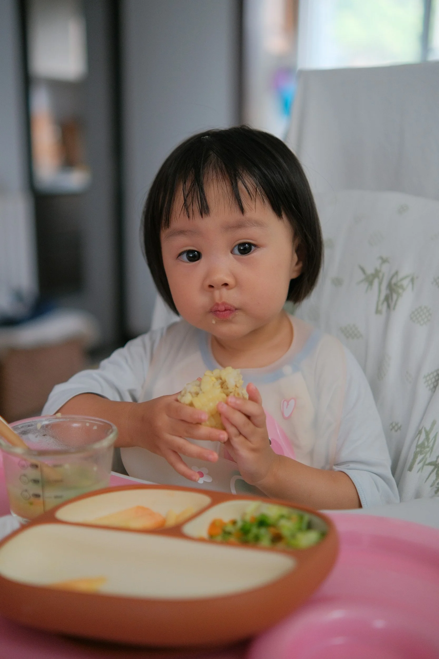 A young Asian girl with short black hair sitting at a table, holding and eating a piece of food with a thoughtful expression, in a cozy, well-lit room with a window.